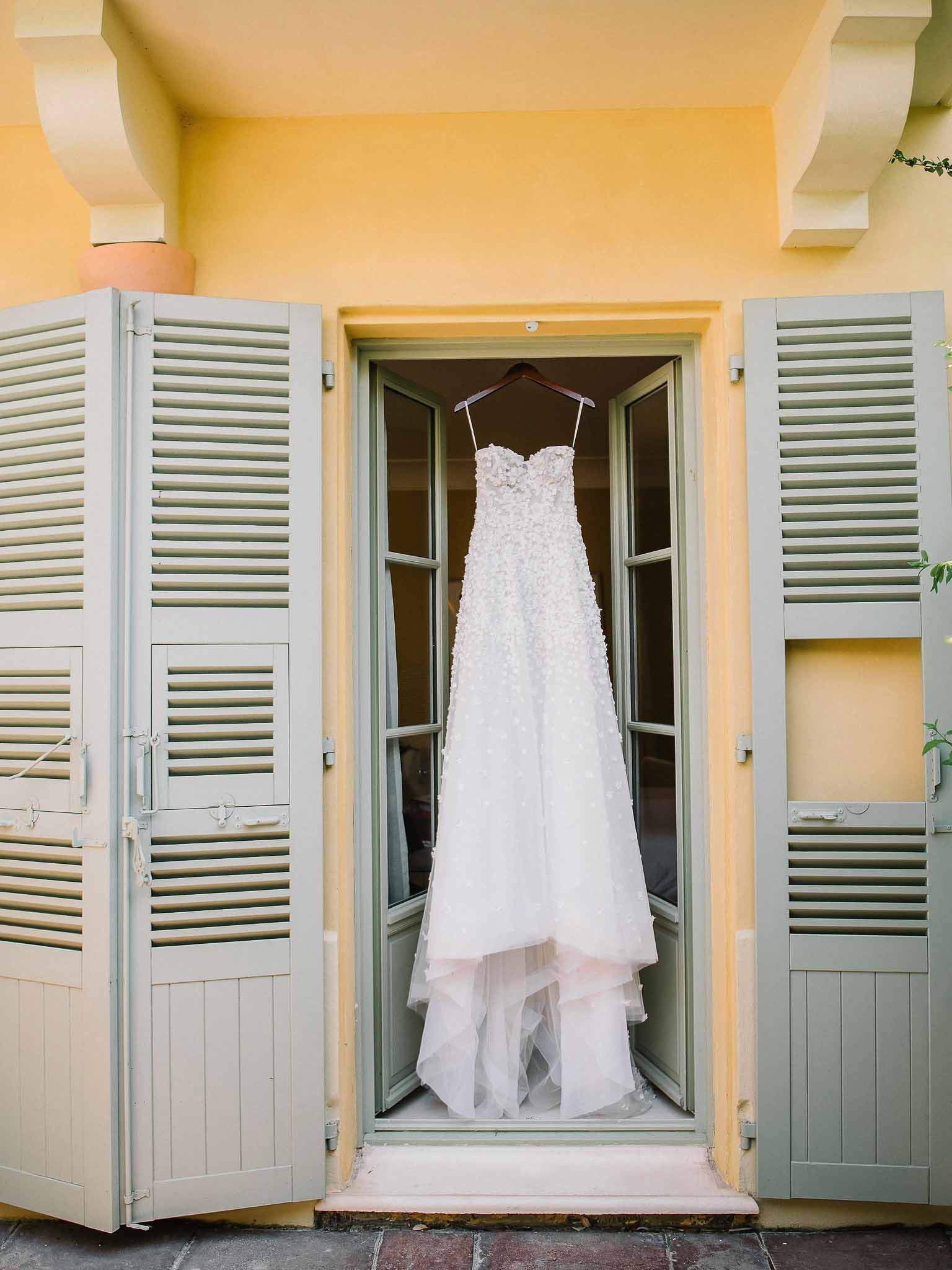 Ivory wedding dress with beaded bodice hanging on wooden hanger framed by sage shutters