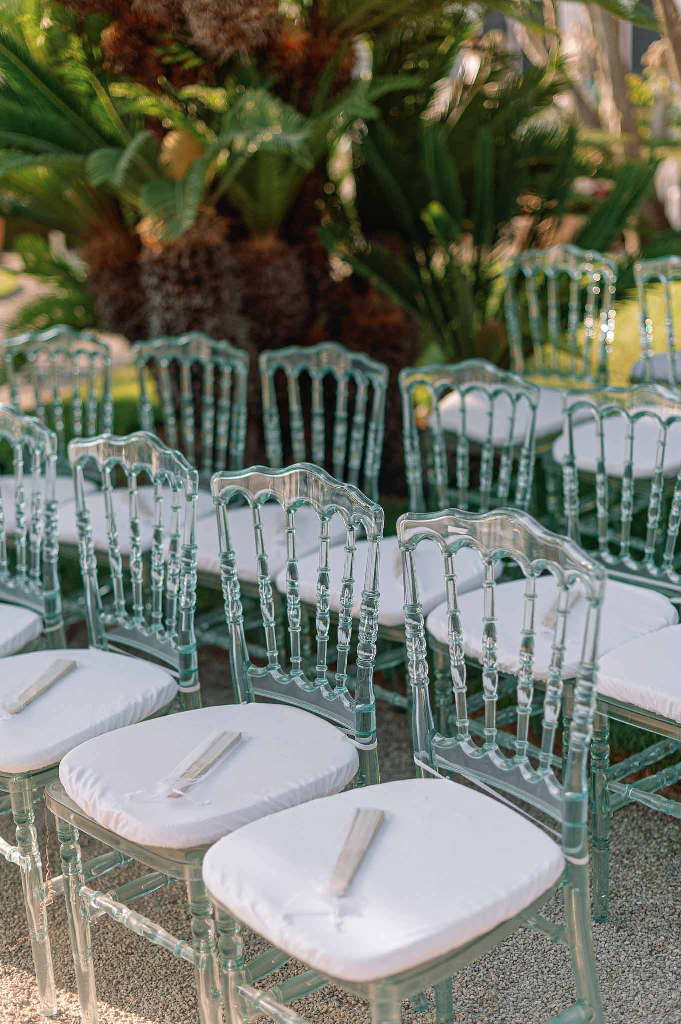Outdoor ceremony seating with acrylic chairs and white cushions in garden setting with lush greenery