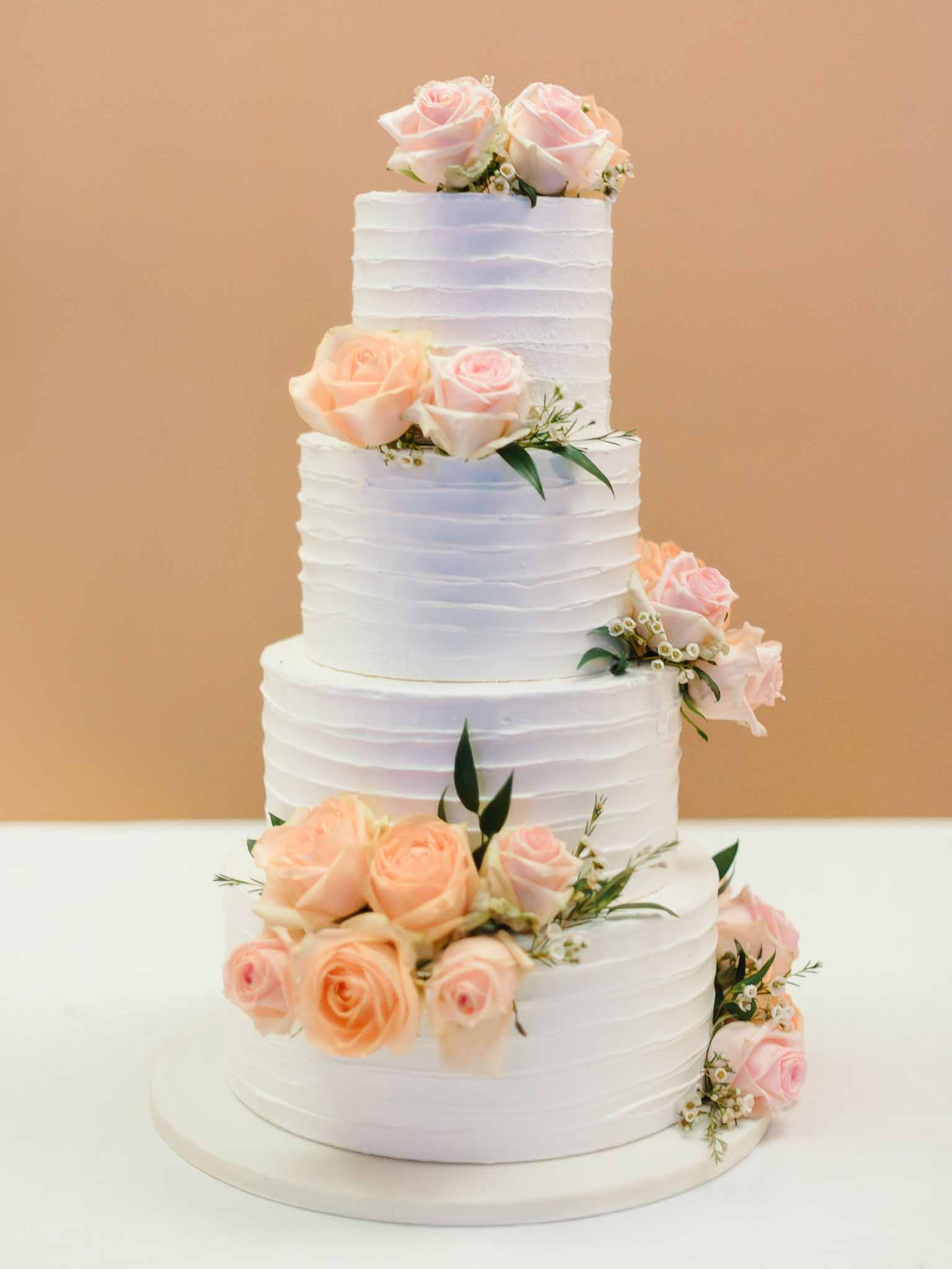 Four-tier wedding cake with cascading peach and pink roses against neutral backdrop