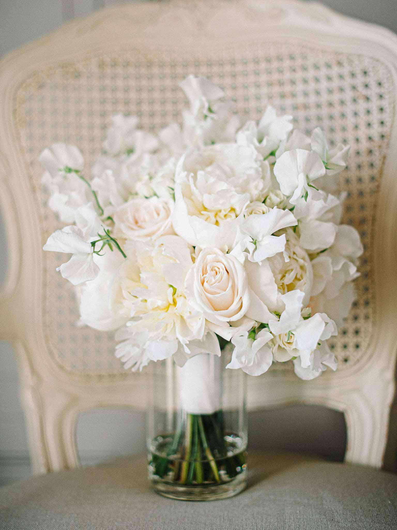 White peony and blush rose bridal bouquet in glass vase on neutral surface