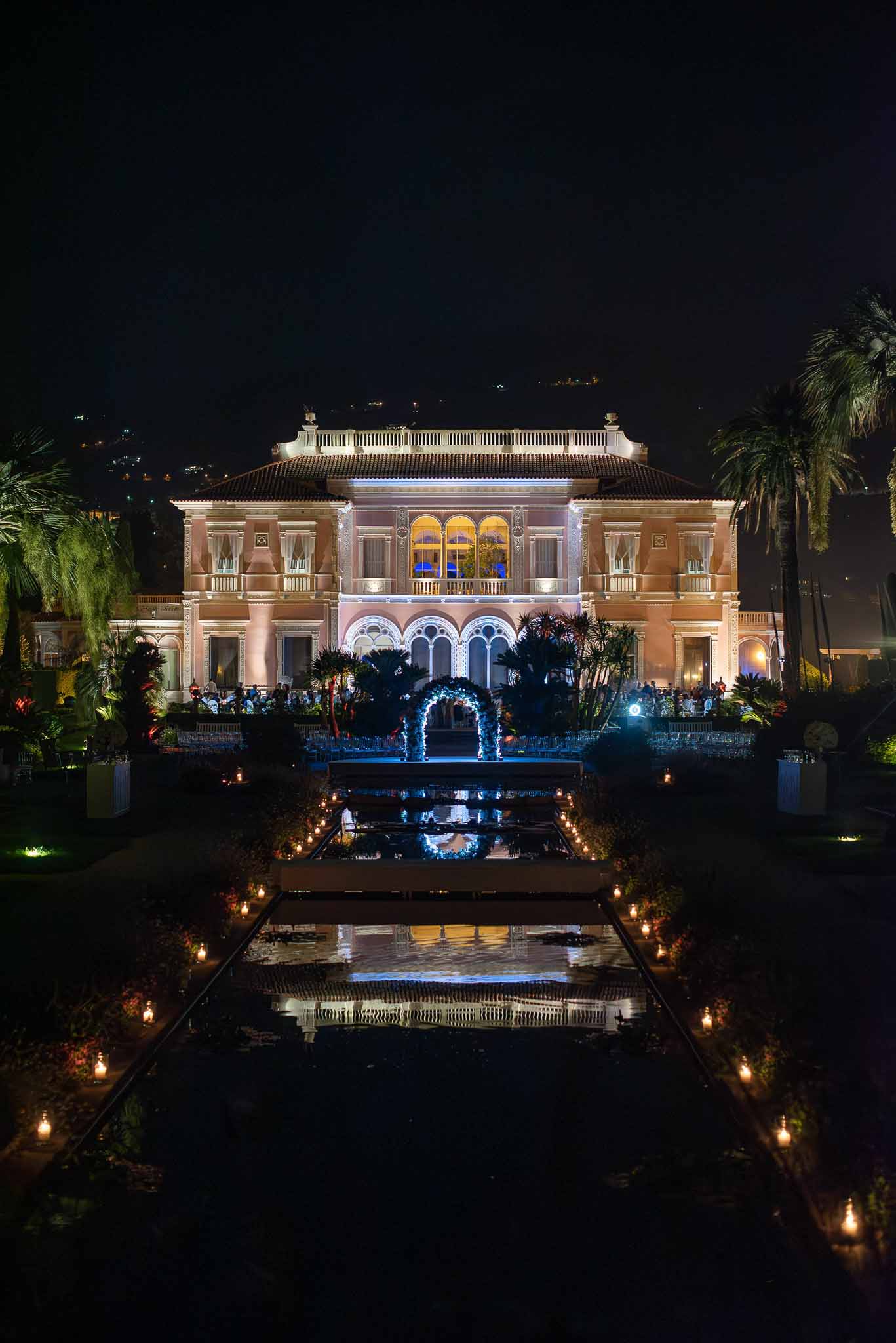 Illuminated neoclassical villa at night with wedding guests on terrace and formal gardens