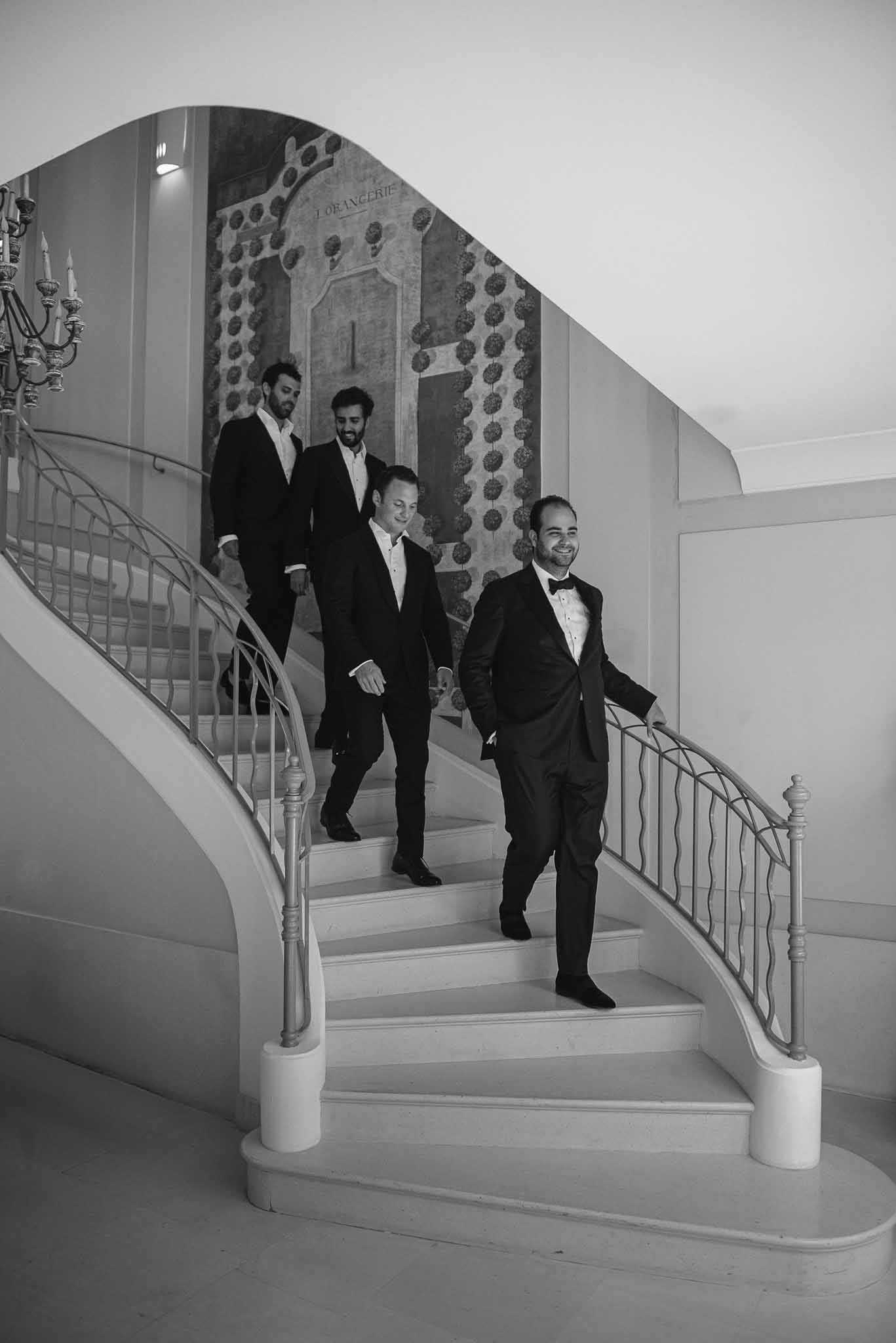 Four groomsmen in black tuxedos descending curved staircase at formal indoor wedding venue