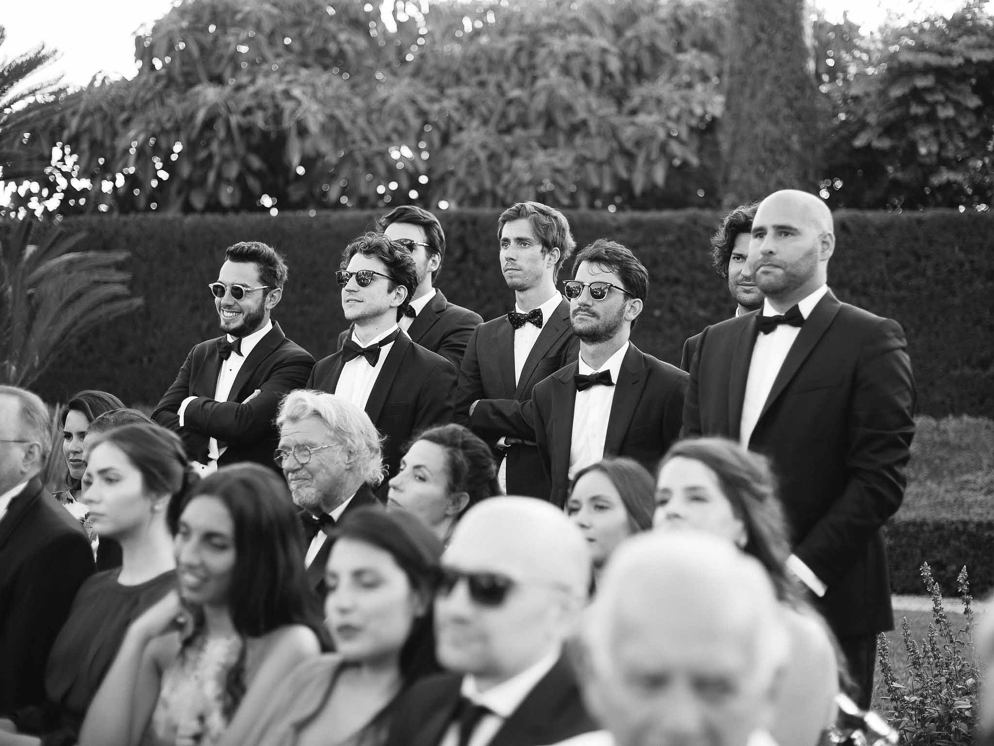 Groomsmen in black tuxedos standing during outdoor wedding ceremony with hedge backdrop