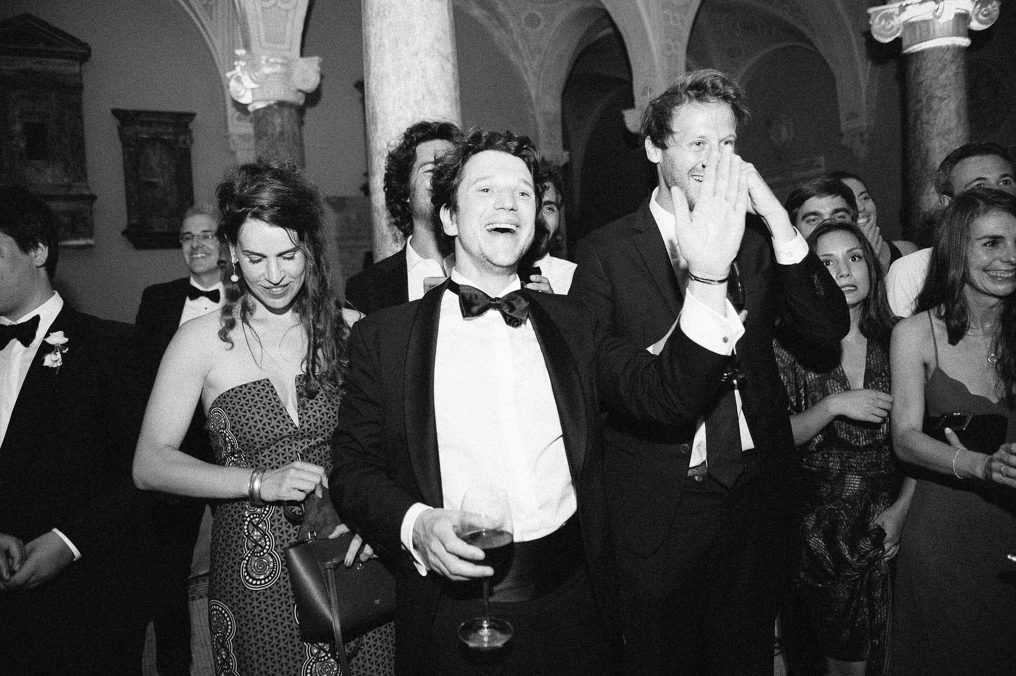 Groom dancing with guests during black-tie reception in classical ballroom with ornate columns
