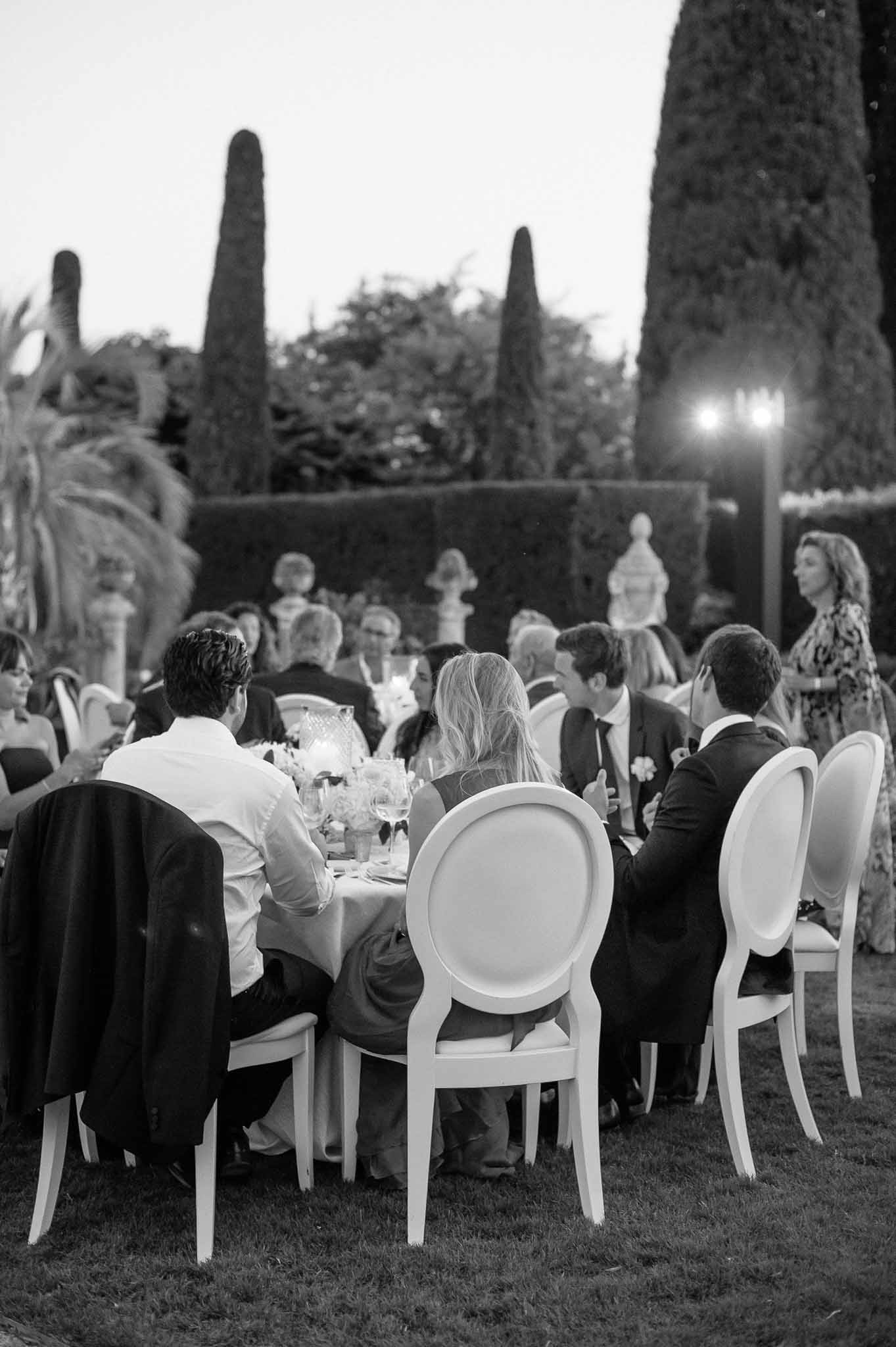 Reception dinner with guests at round tables in formal garden with cypress trees and classical sculptures