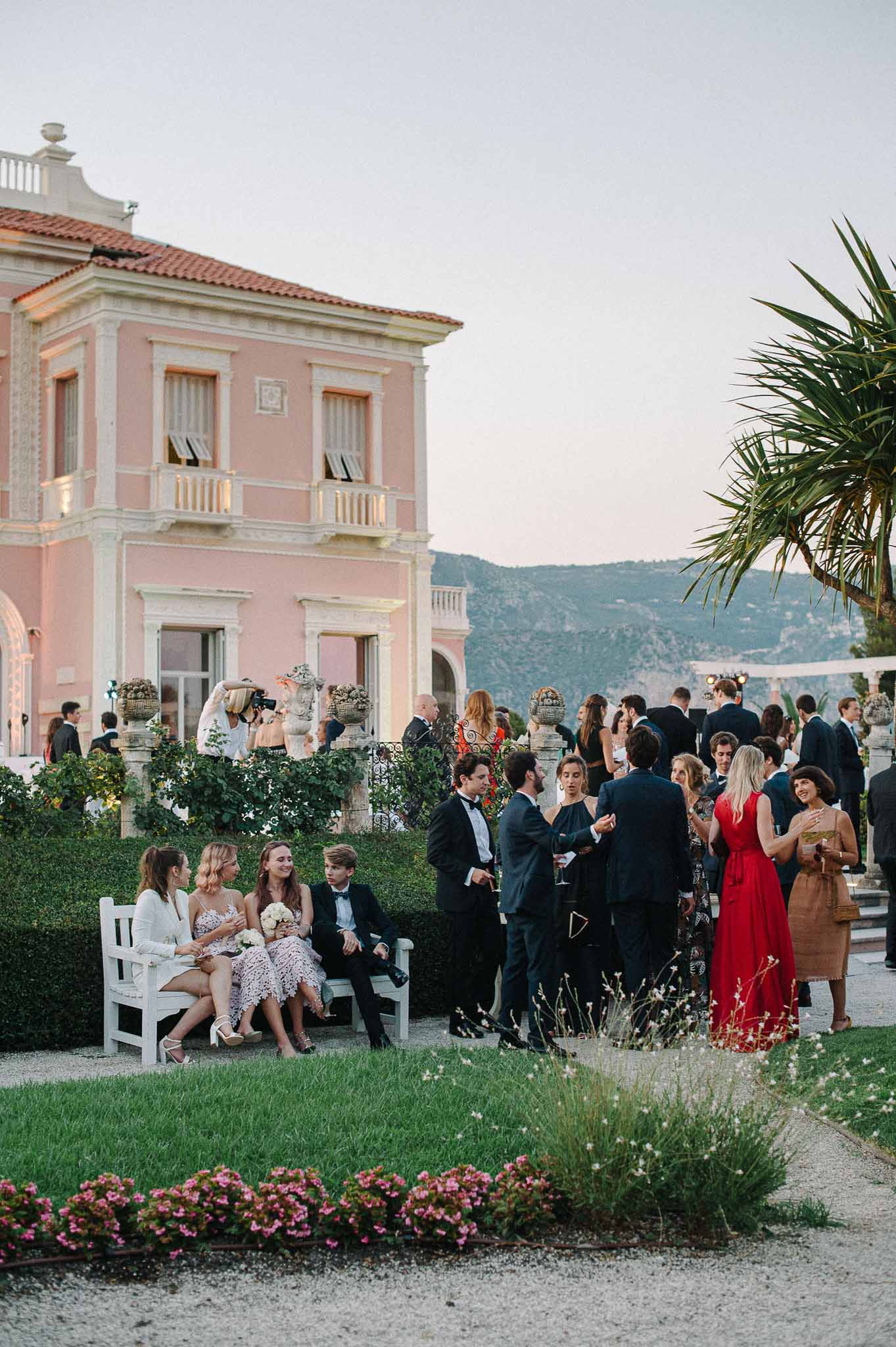 Cocktail hour with guests mingling on terrace of pink neoclassical villa with landscaped grounds