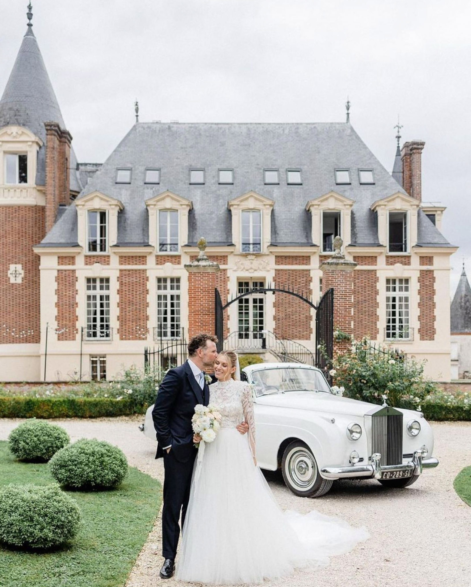 Groom kisses bride on cheek in front of red brick French chateau with vintage white Rolls-Royce