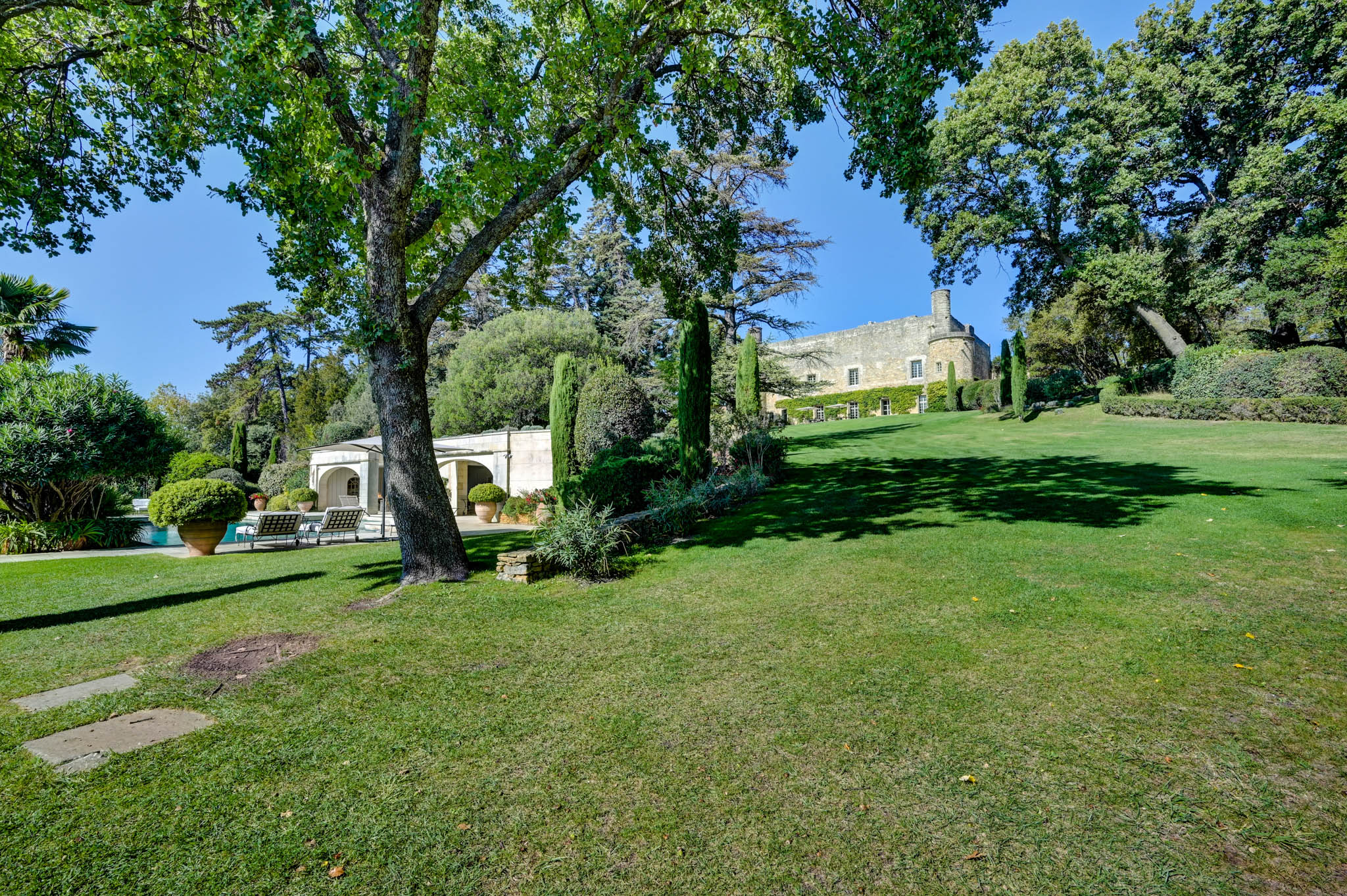 French chateau in pale golden stone with round tower, formal gardens, cypress trees, and white arched pavilion