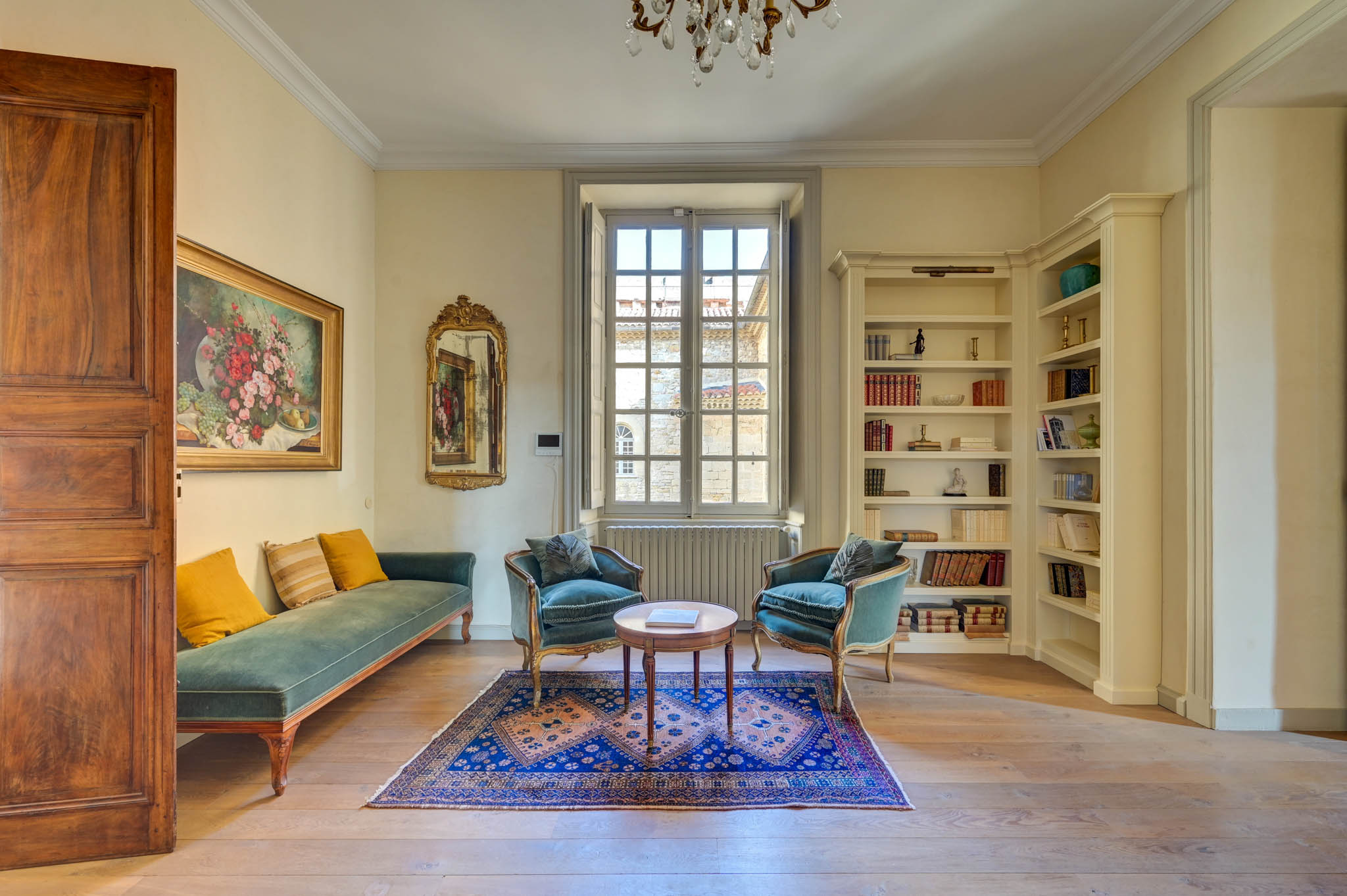French chateau salon with green velvet daybed, teal armchairs, gilt chandelier, and built-in bookcase