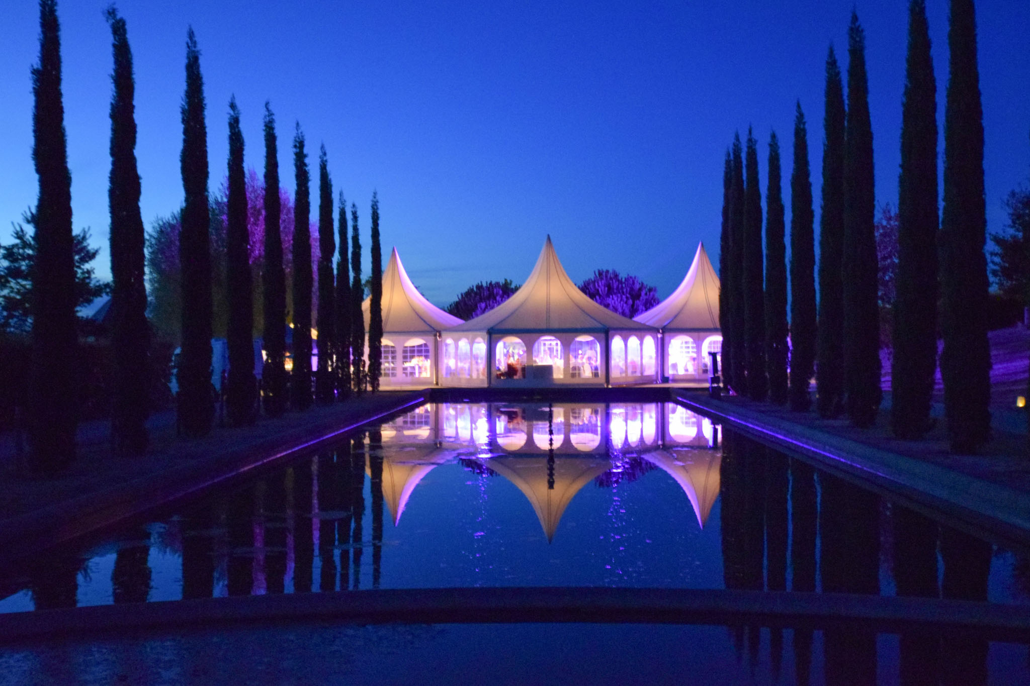 White marquee tent lit from within at dusk reflected in ornamental pool flanked by cypress trees with purple uplighting