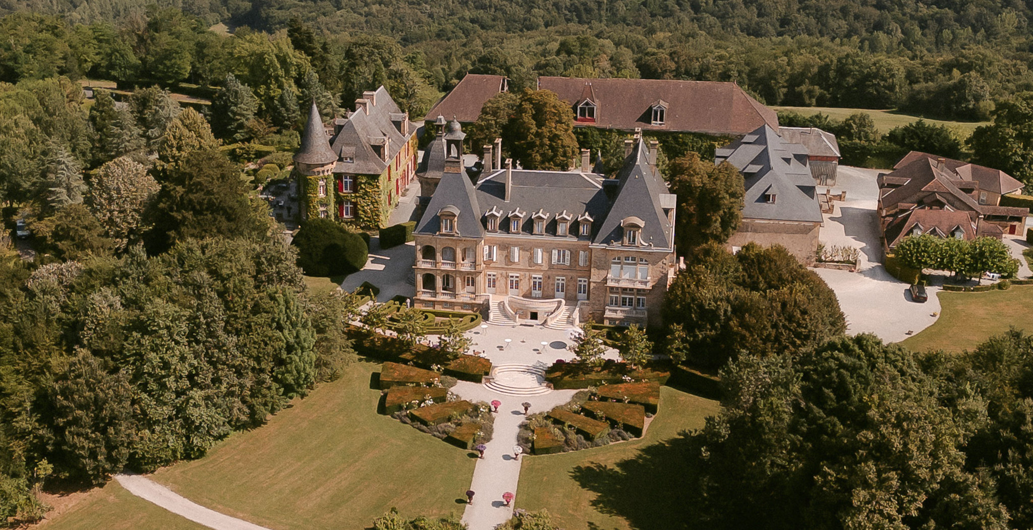 Aerial drone view of French chateau estate with slate mansard roofs, formal geometric gardens, and woodland surroundings