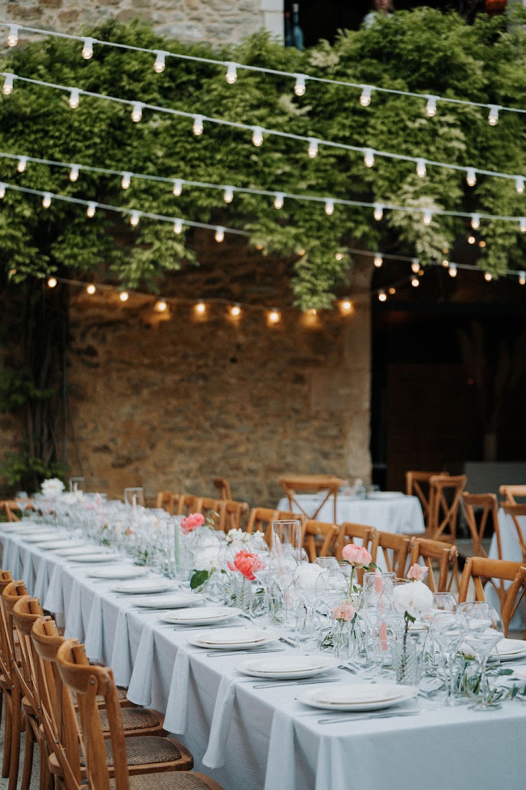 Long reception table with blue-grey linen, coral peonies, and string lights in vine-covered stone courtyard