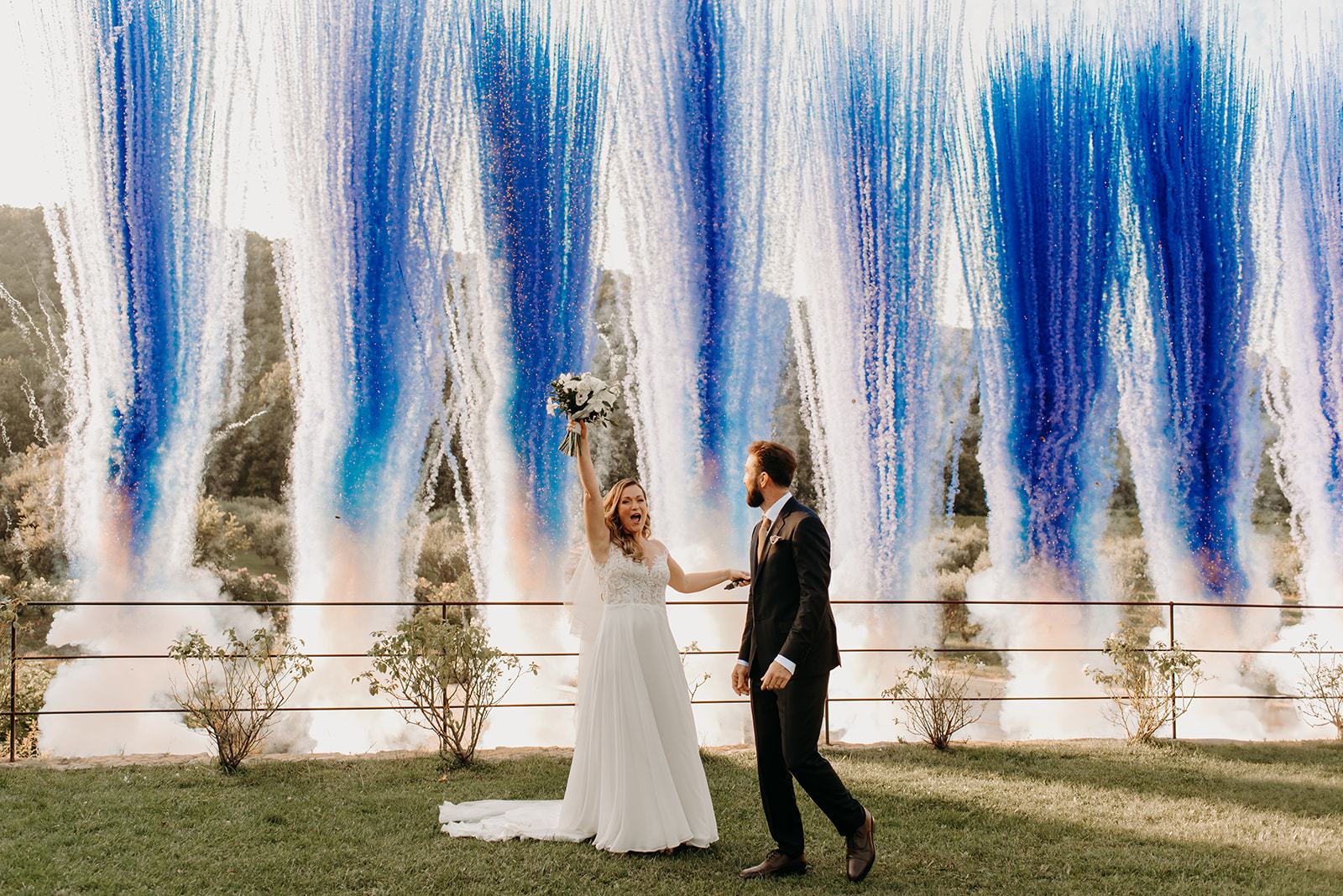 Couple walking through blue and silver pyrotechnic fountains on lawn bride raising bouquet in celebration