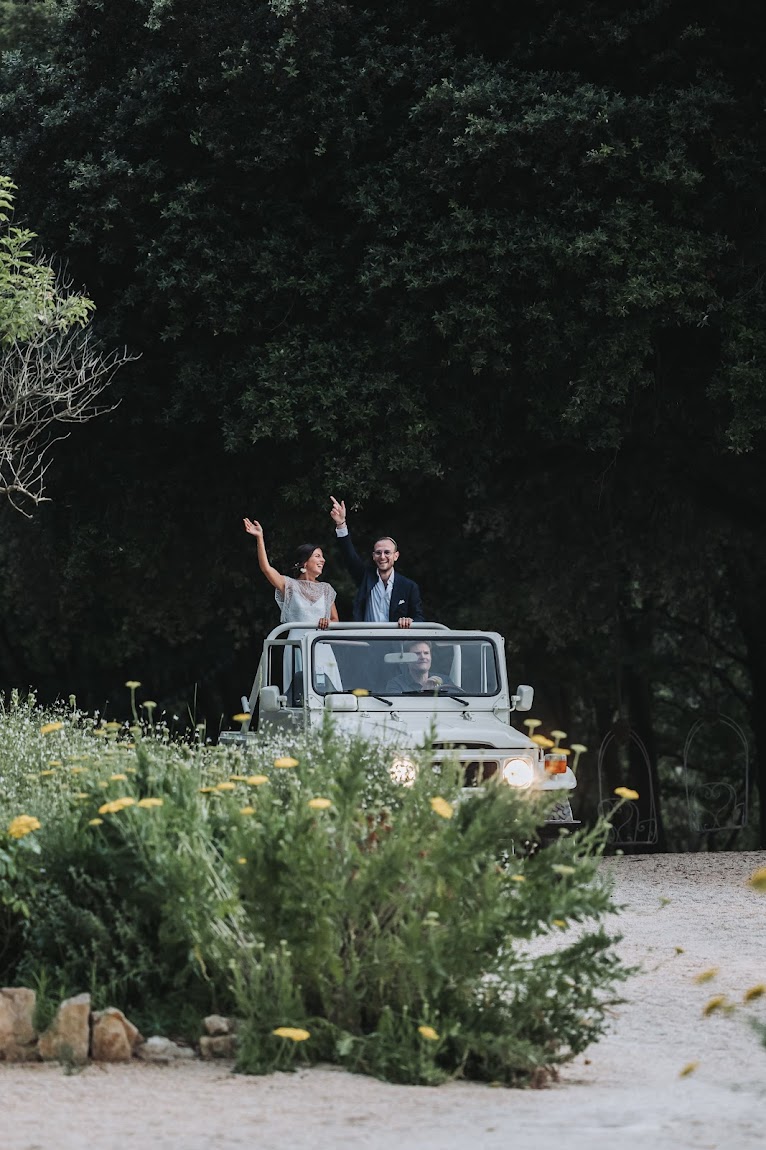 Couple waving from open-roof vintage 4x4 on gravel driveway with wild yellow flowers and trees at dusk