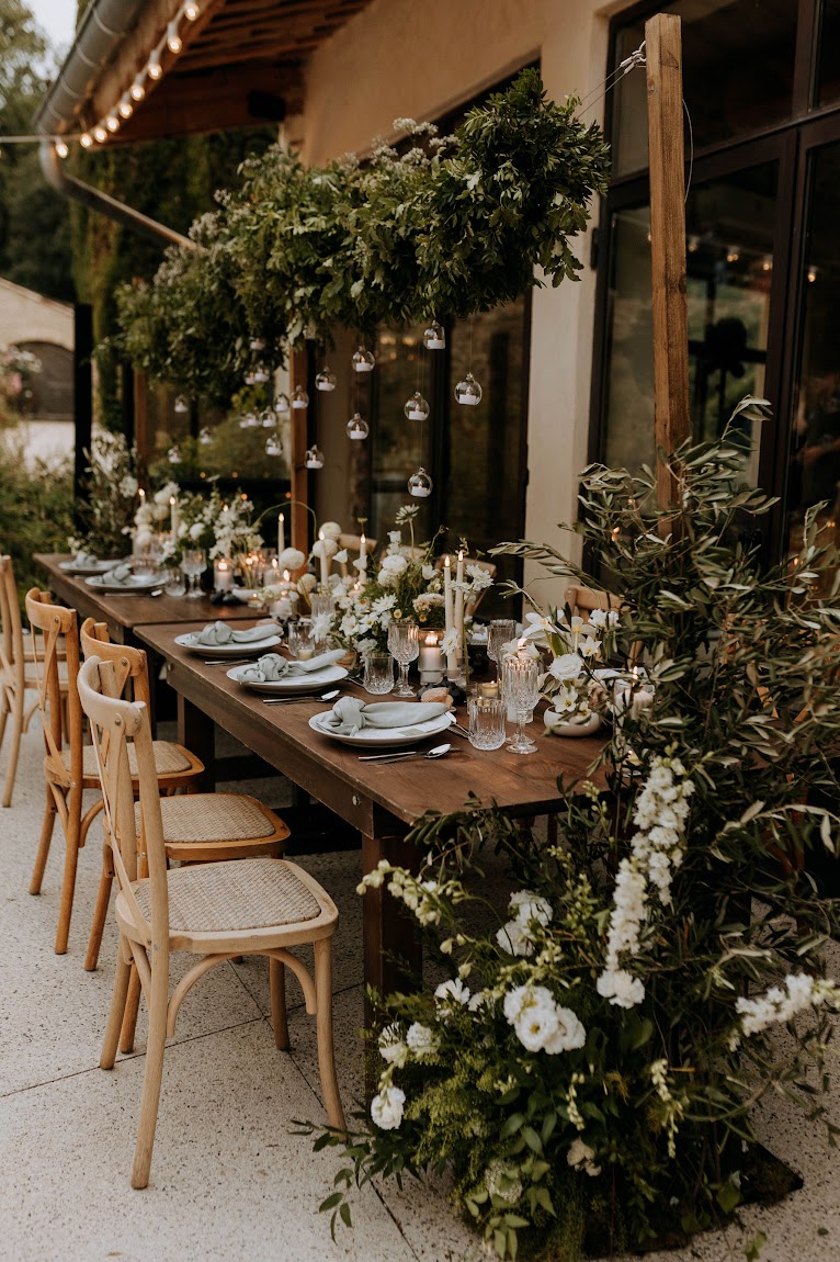 Farm table with white ranunculus foliage runner and hanging greenery installation on covered terrace