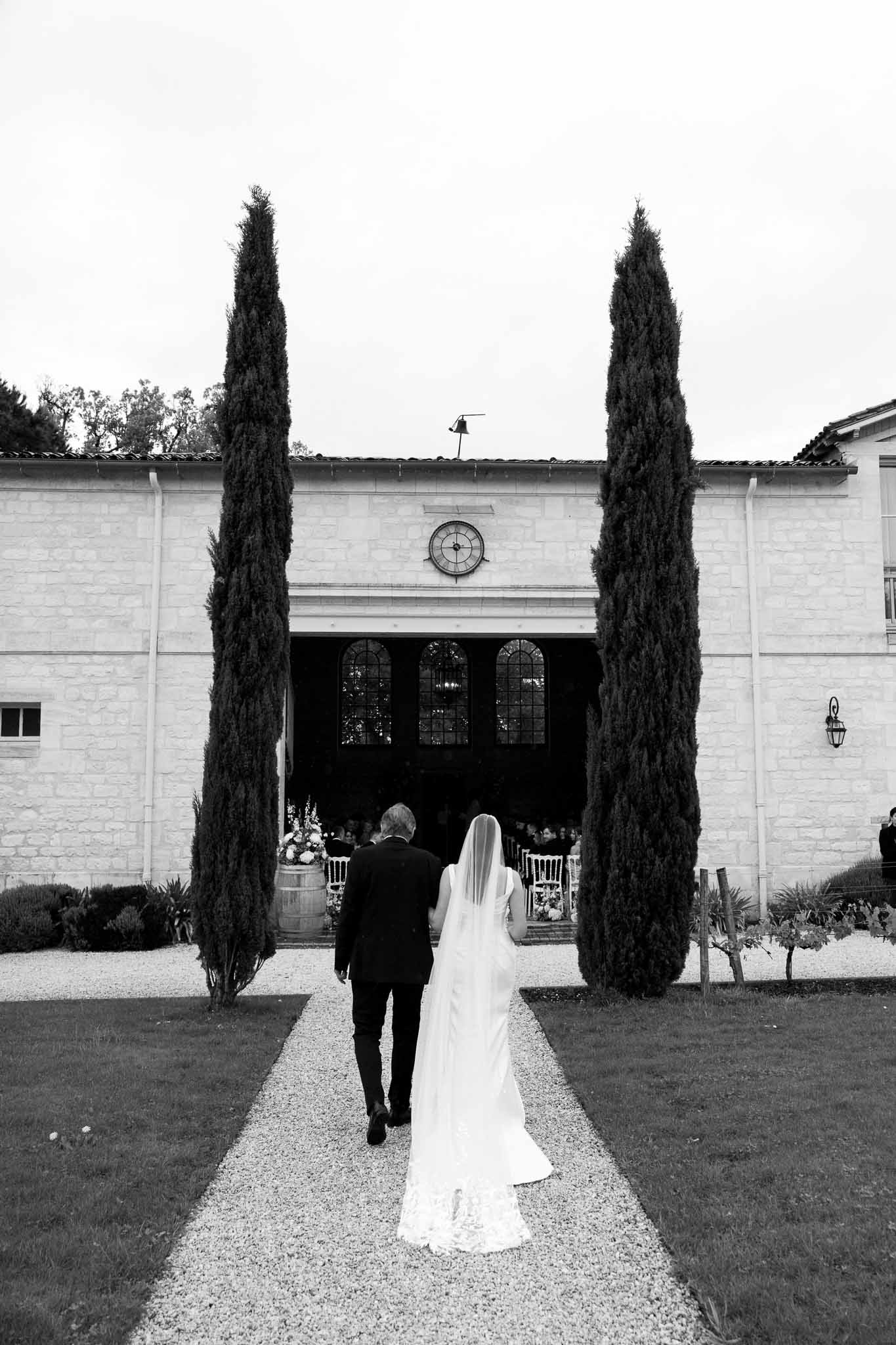 Bride and groom walking toward white chapel with cypress trees during wedding ceremony processional