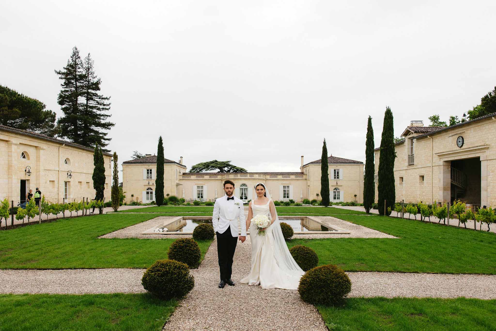Bride and groom hold hands on gravel path between reflecting pools in formal chateau gardens with cypress trees