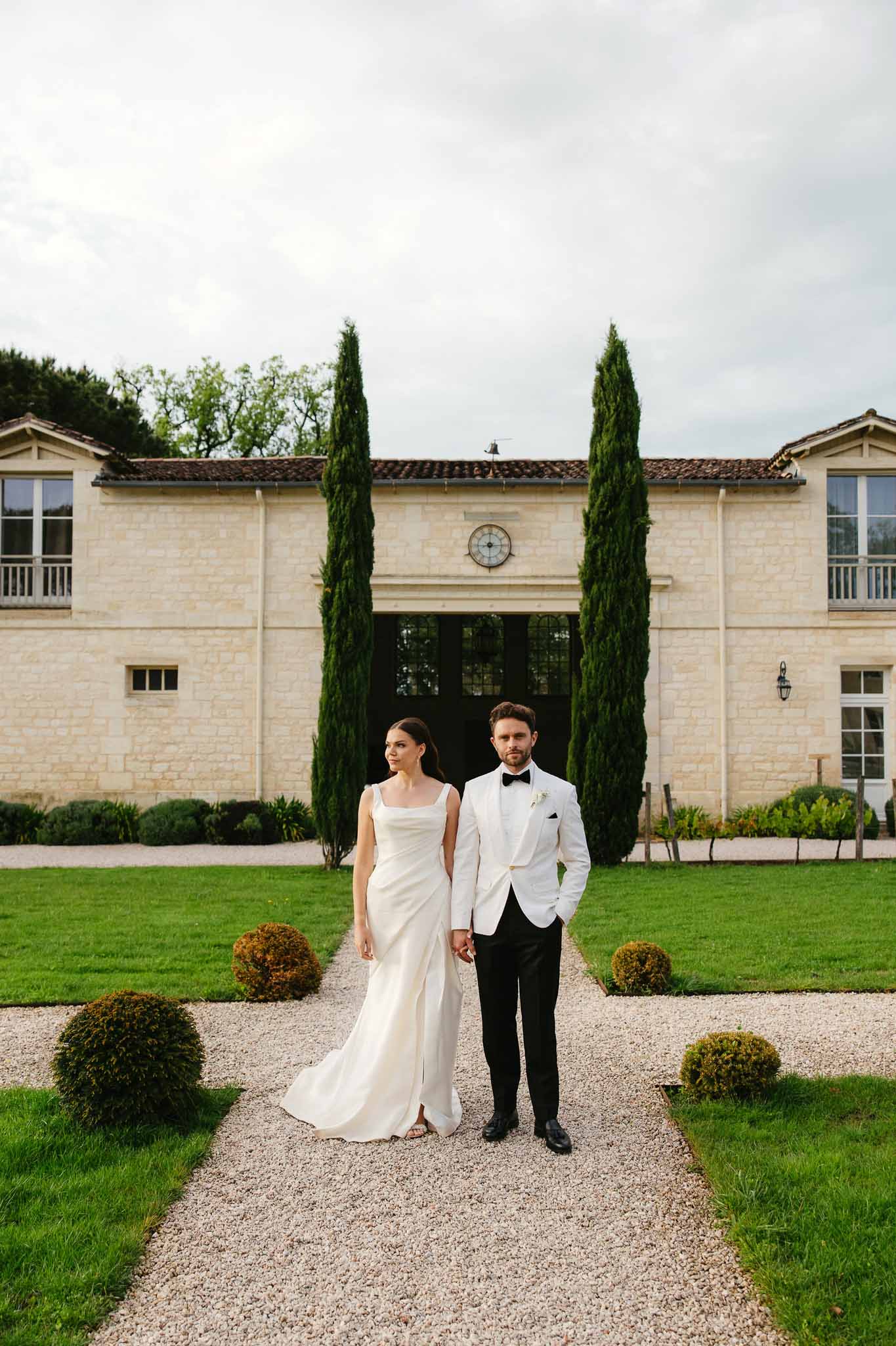 Bride and groom portrait on pathway to cream stone mansion with cypress trees