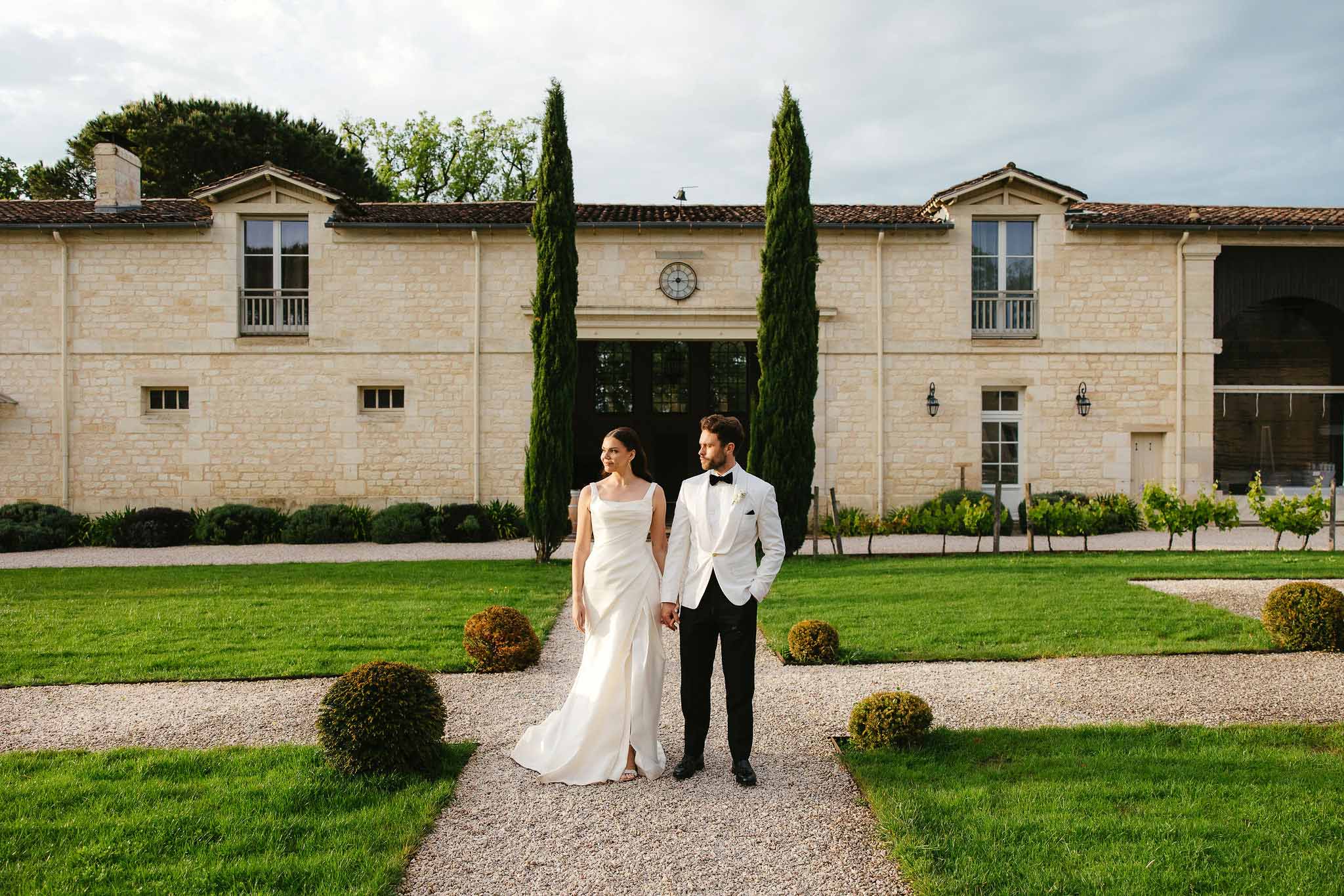 Bride and groom posing together in front of stone manor house with formal gardens