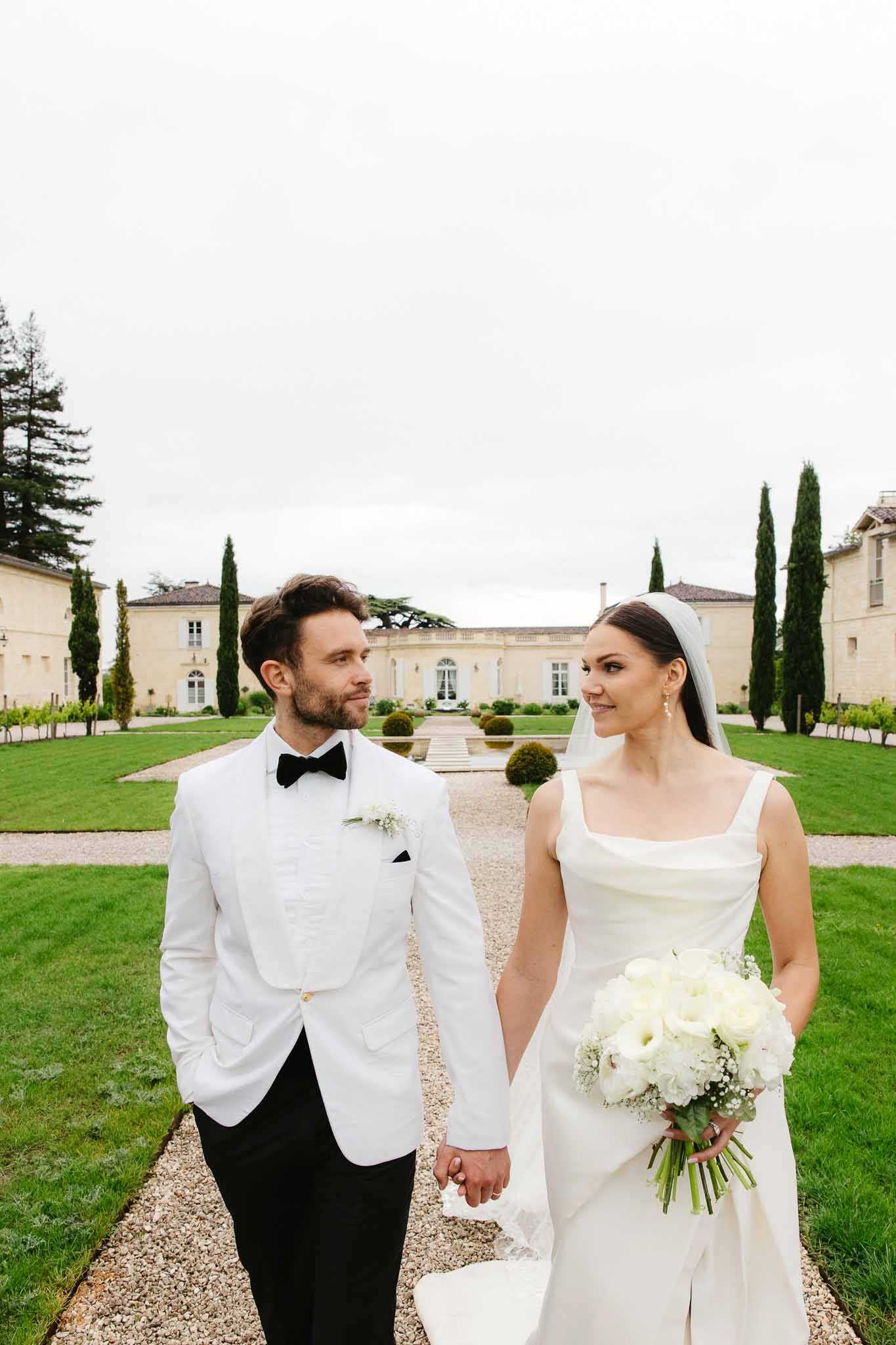 Bride and groom walking hand-in-hand along gravel pathway at French chateau estate with manicured gardens