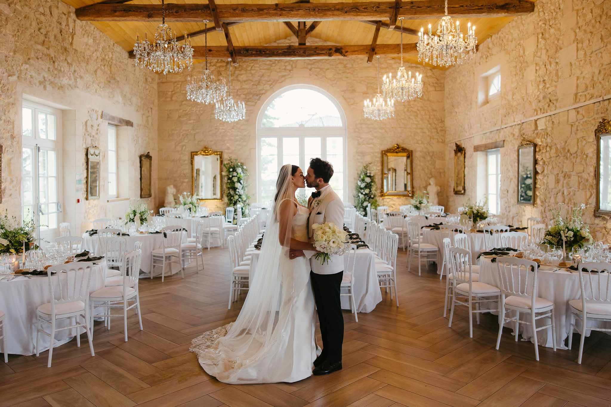 Bride and groom kissing in a chandelier-lit chateau reception hall with white-dressed tables and green napkins