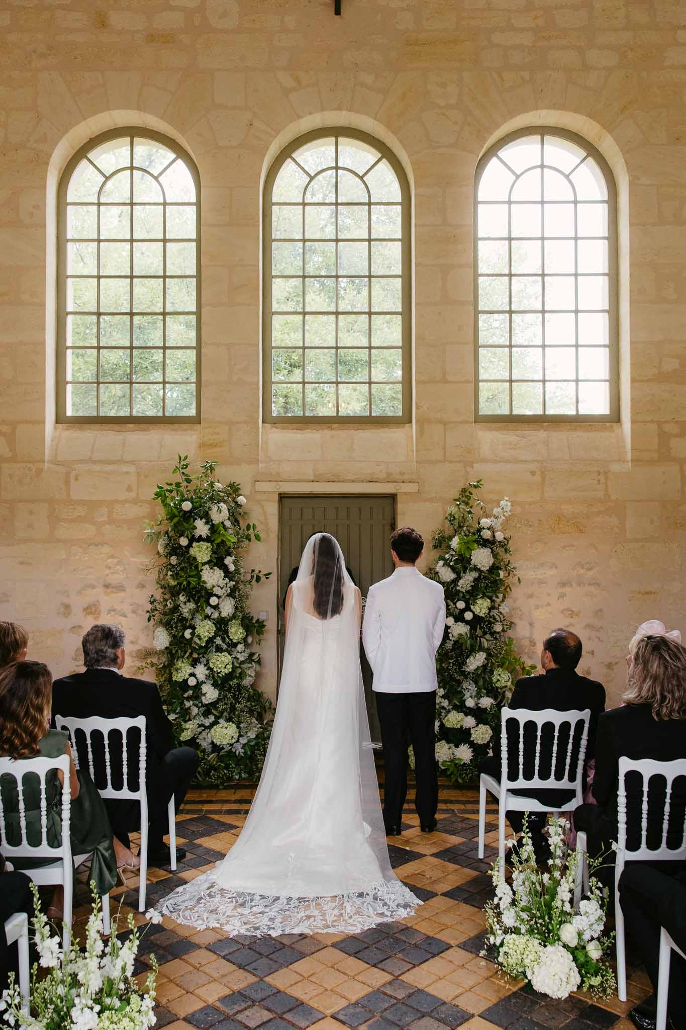 Wedding ceremony in stone chapel with arched windows and floral arrangements