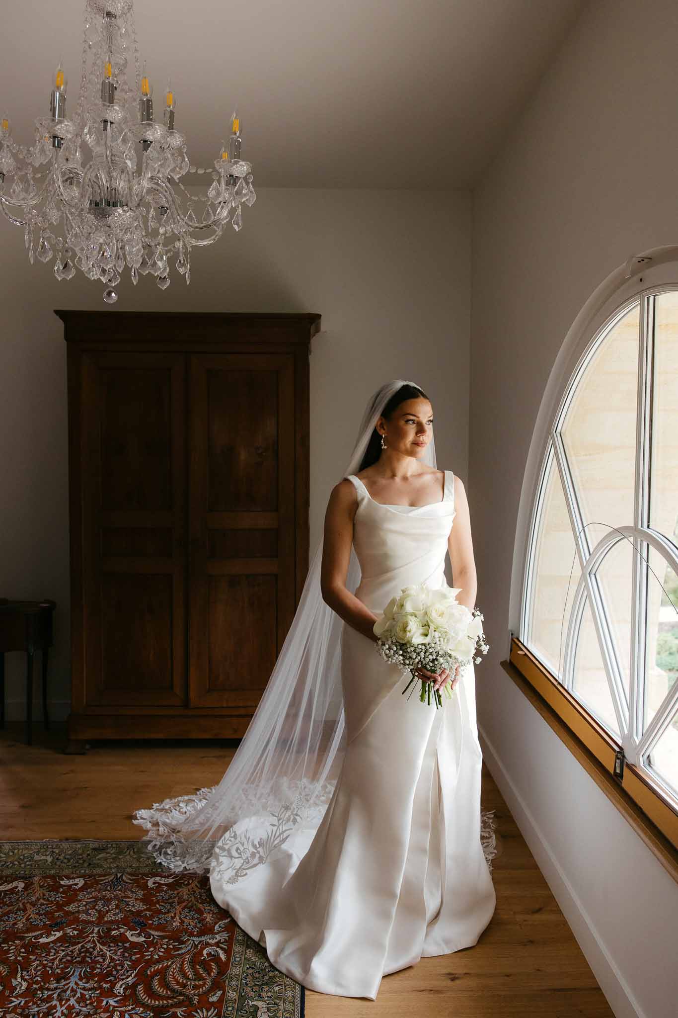Bride in ivory satin gown with tulle veil holding white rose bouquet in elegant interior space with chandelier