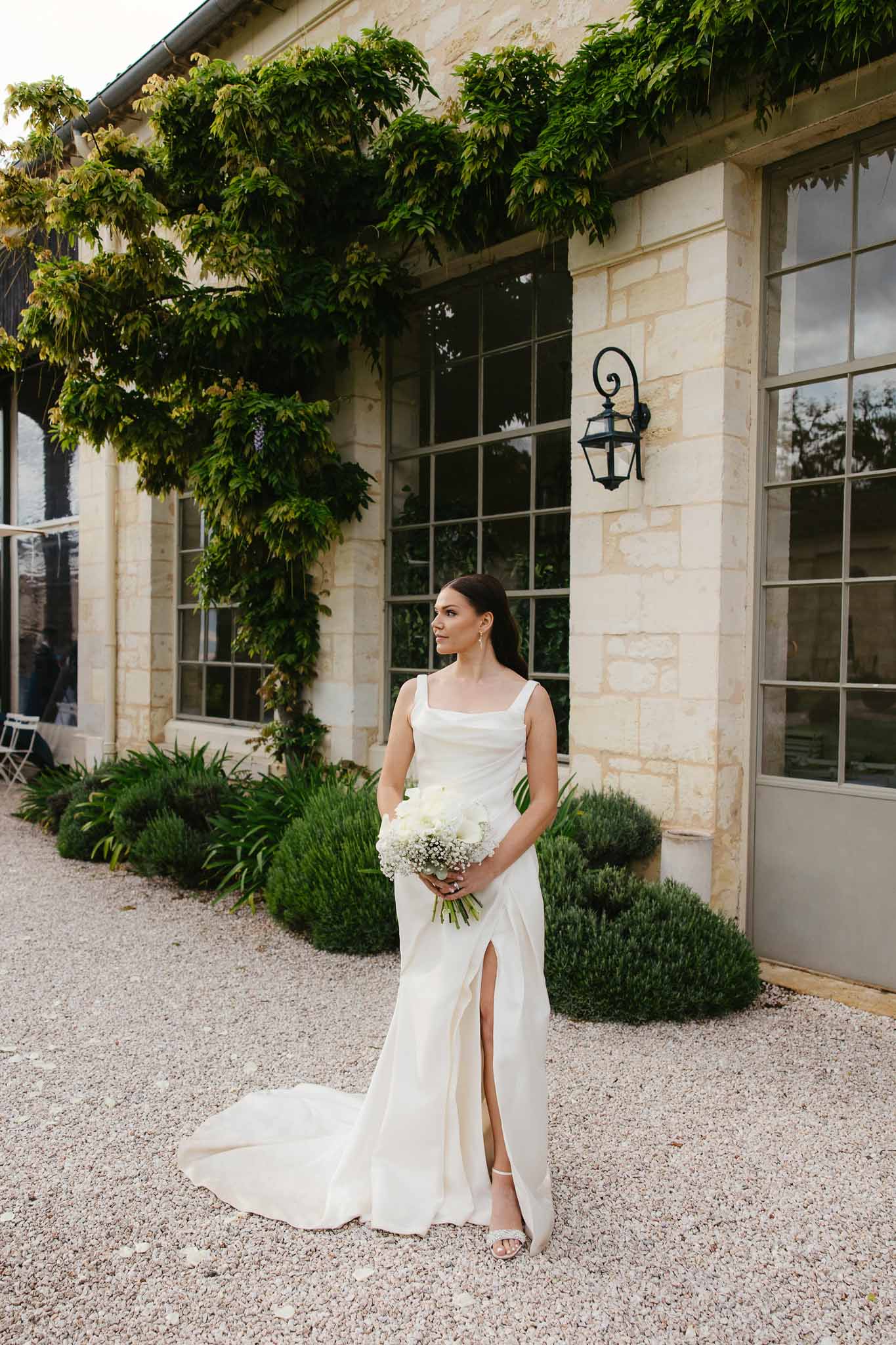 Bride in ivory sleeveless square-neck gown holding white rose bouquet on gravel courtyard of limestone chateau