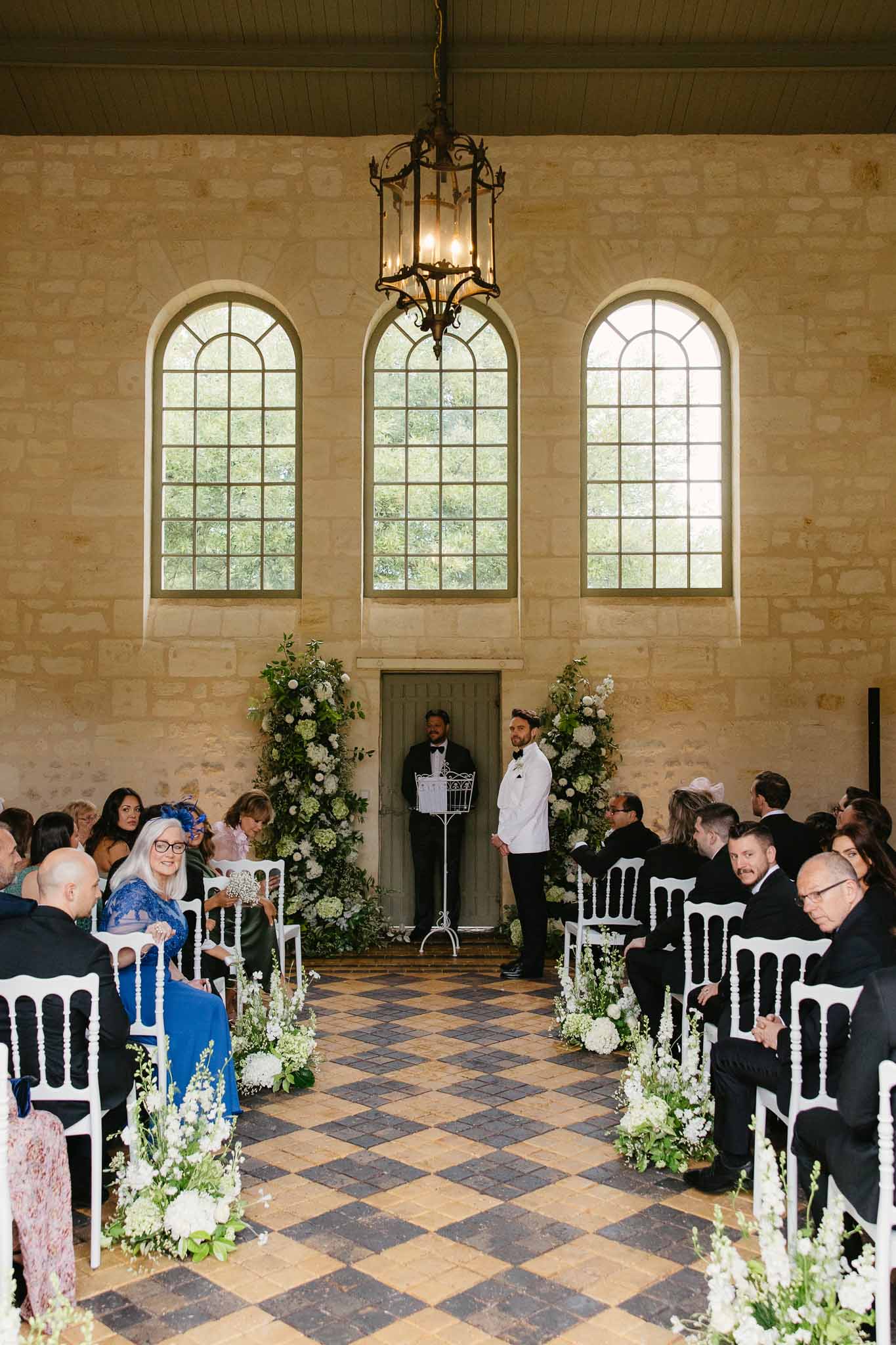 Wedding ceremony in stone chapel with arched windows and white floral arrangements