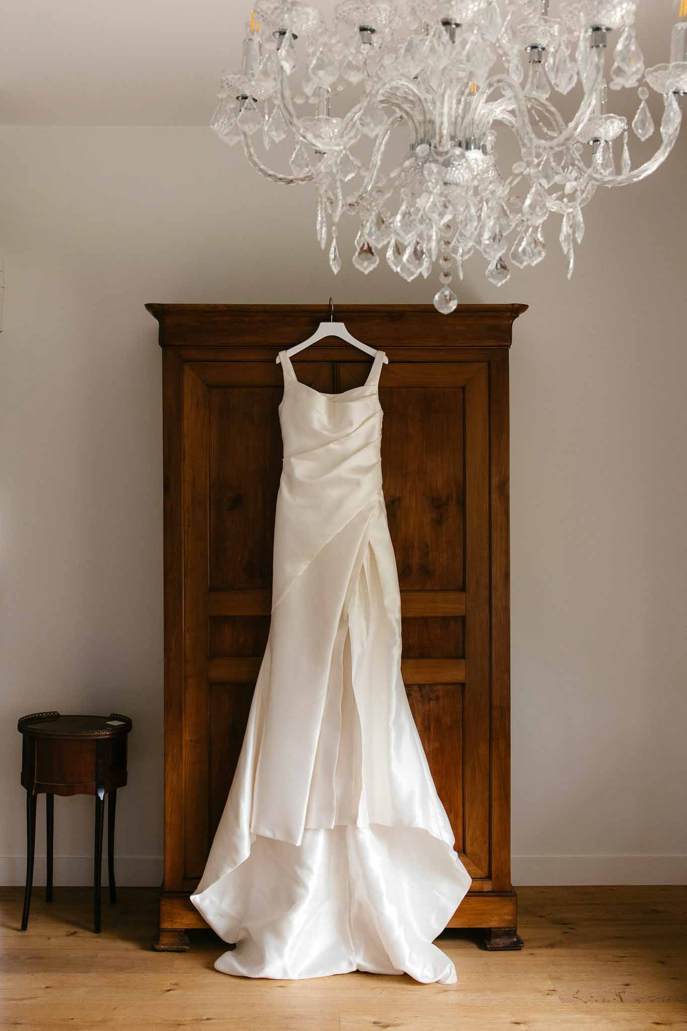 Ivory silk wedding dress hanging on antique armoire in bridal suite with crystal chandelier