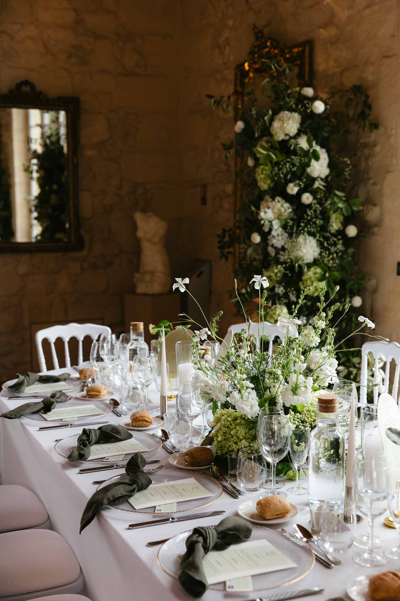 Reception table with gold-rimmed chargers, olive napkins, white hydrangea centerpiece and floral installation