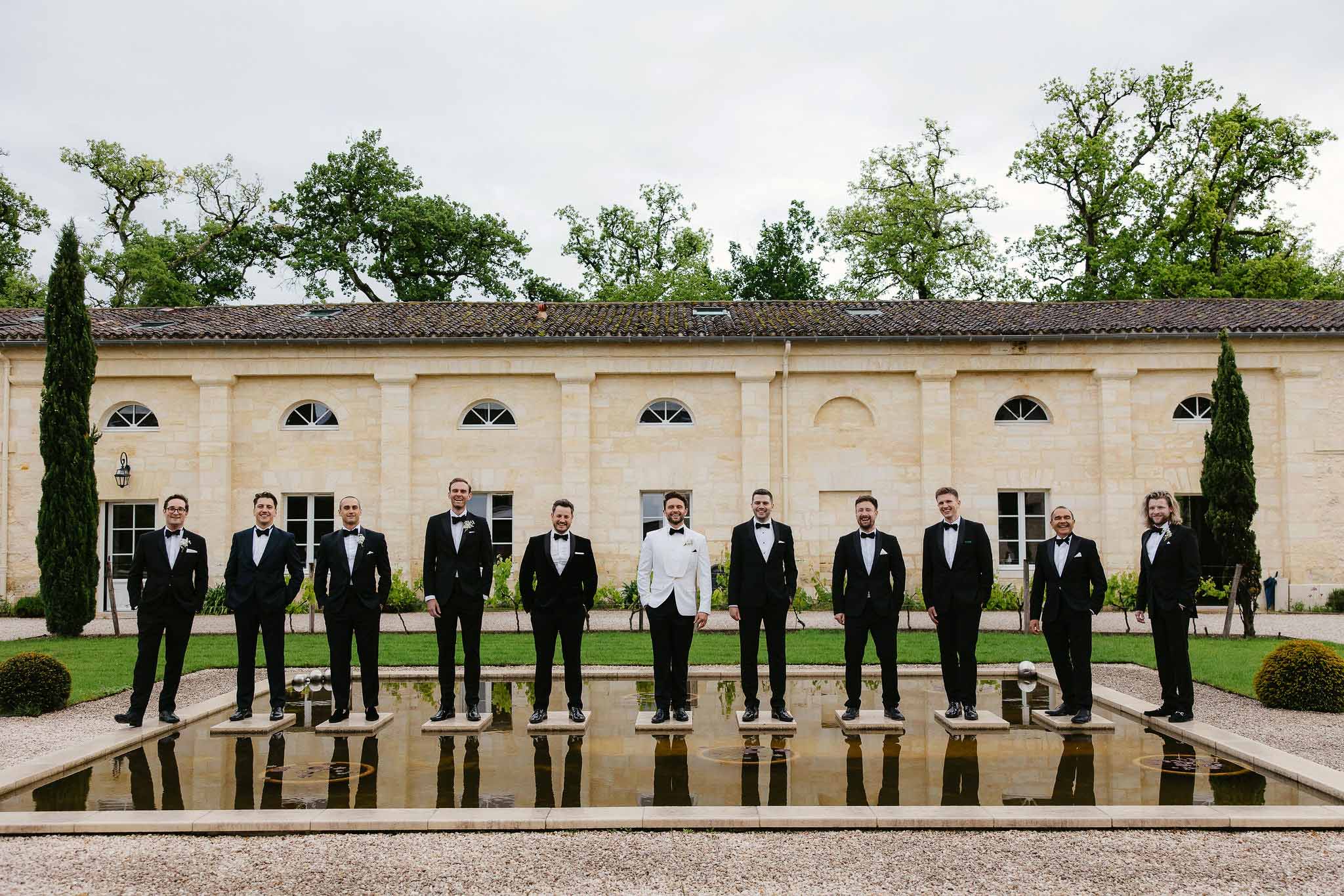 Groom and eleven groomsmen in formal tuxedos standing by reflecting pool at classical stone venue