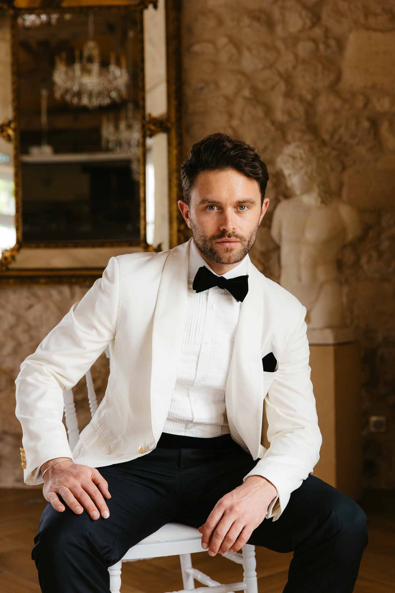 Groom in ivory shawl-lapel dinner jacket seated in chateau salon with gilt mirror and bust