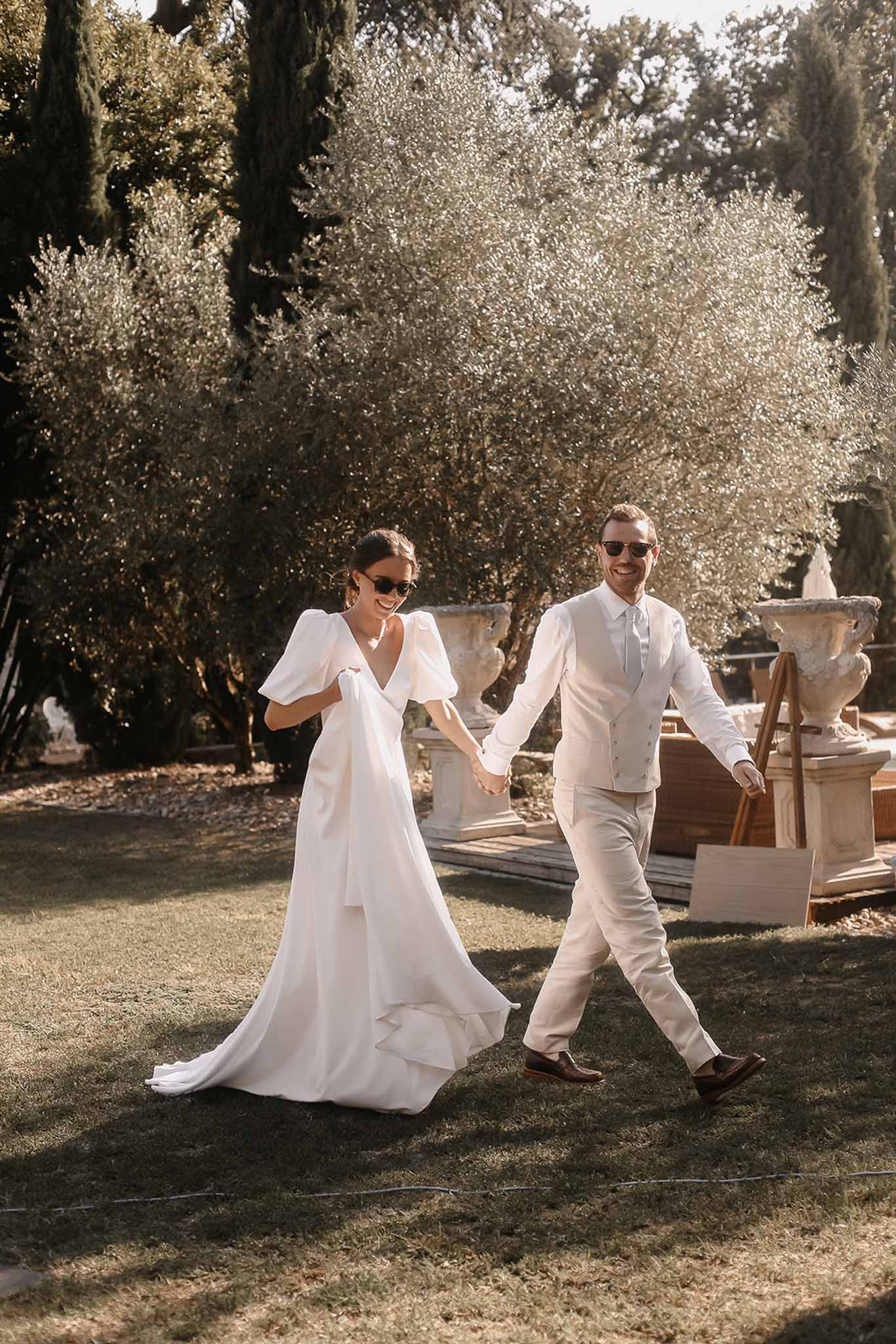Bride and groom walking hand-in-hand in Mediterranean villa garden with cypress trees and flowering shrubs