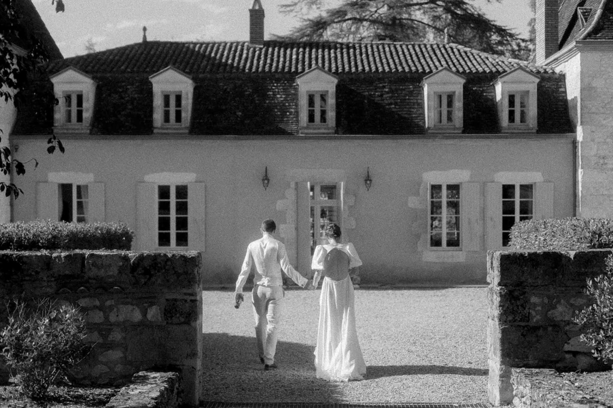 Bride and groom walking toward classical stone manor house with mansard roof and formal courtyard