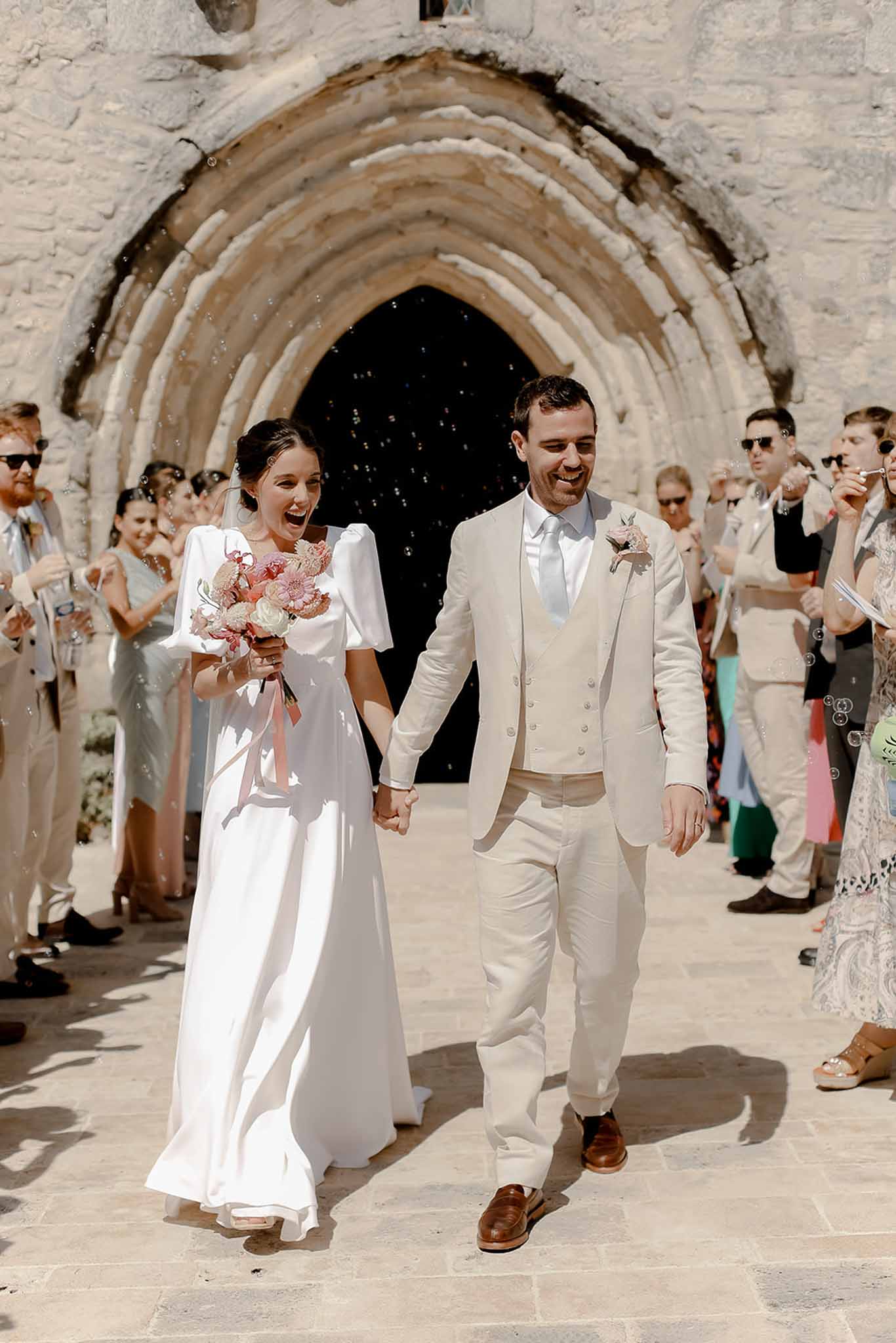 Bride and groom walking through stone archway during recessional with guests blowing bubbles at outdoor ceremony