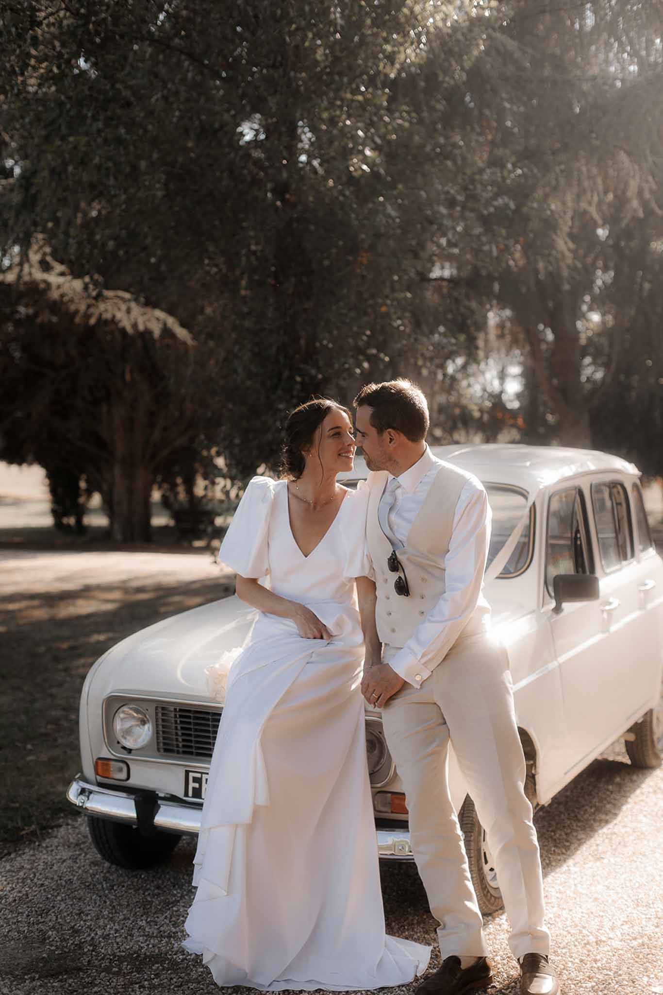 Bride and groom sitting on vintage cream car along tree-lined avenue in warm afternoon light