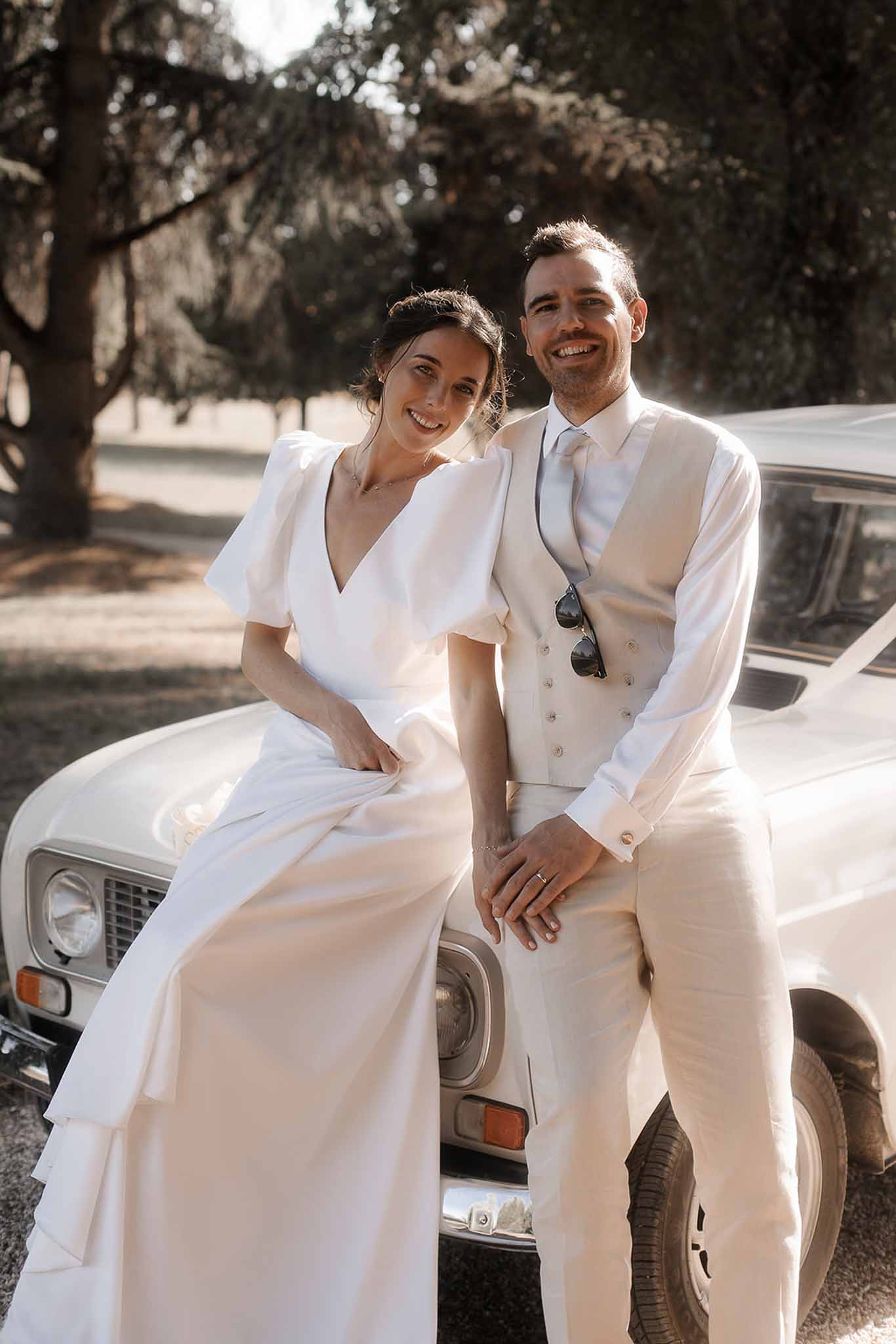 Bride and groom posing on vintage white car in tree-lined garden setting