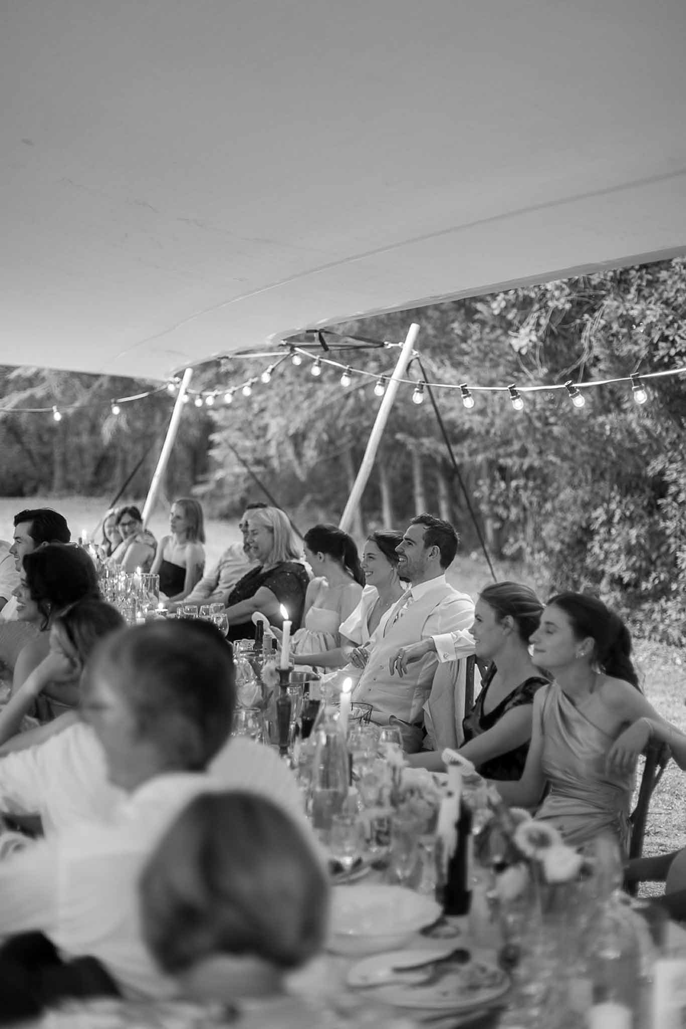 Outdoor reception dinner under marquee tent with guests at long banquet table in garden setting