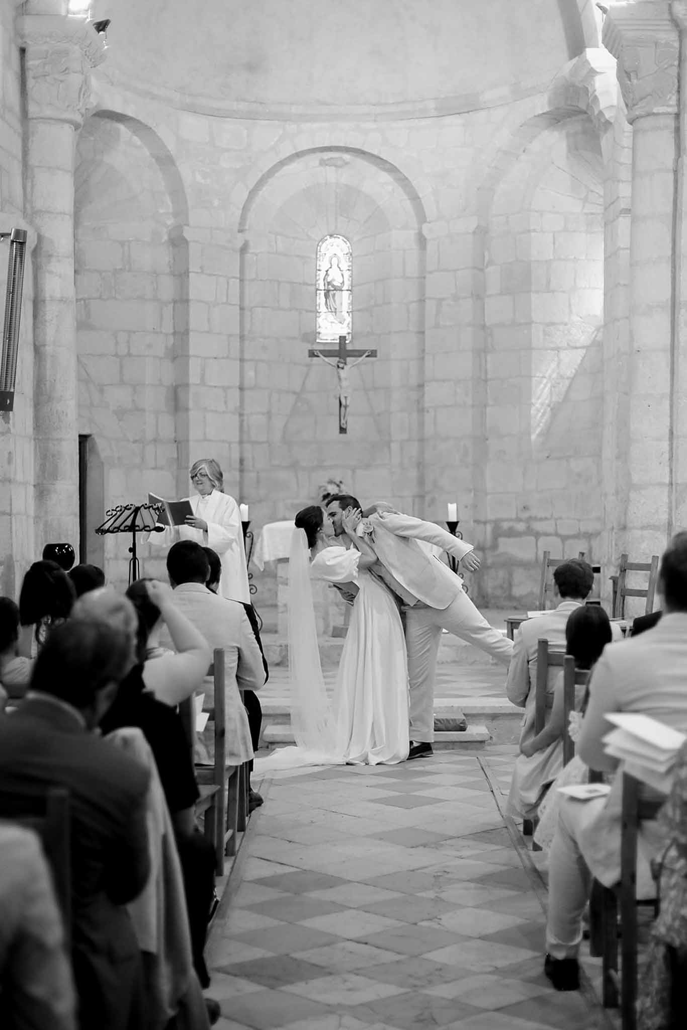 Bride and groom kissing during wedding ceremony in Romanesque stone chapel with guests watching