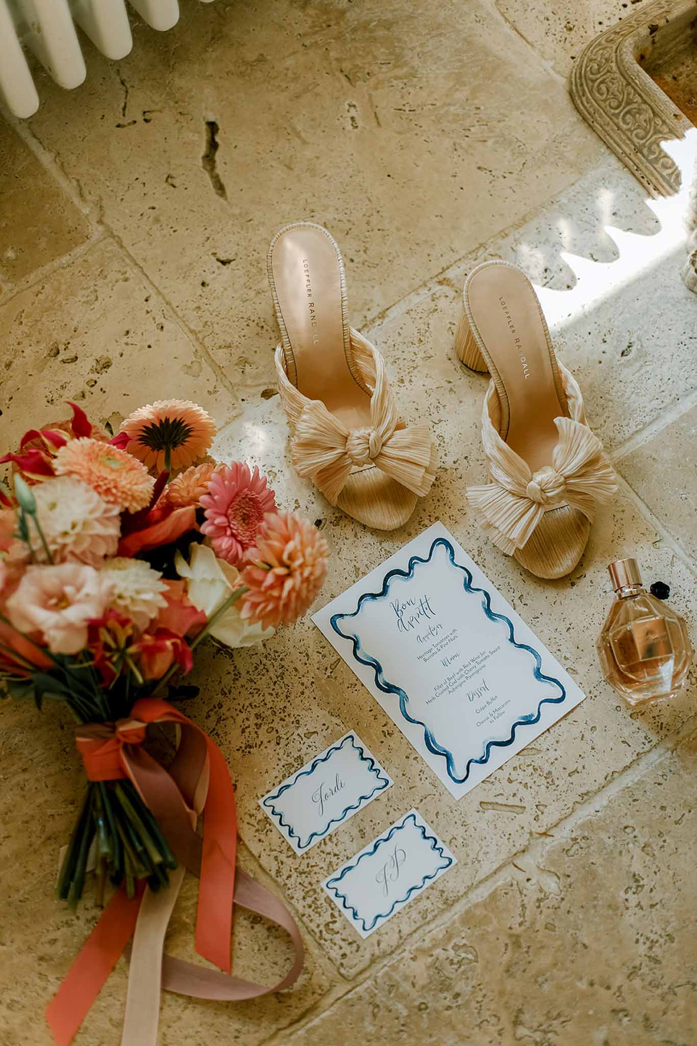Bridal accessories flat lay with coral bouquet, cream shoes, and navy stationery on stone floor