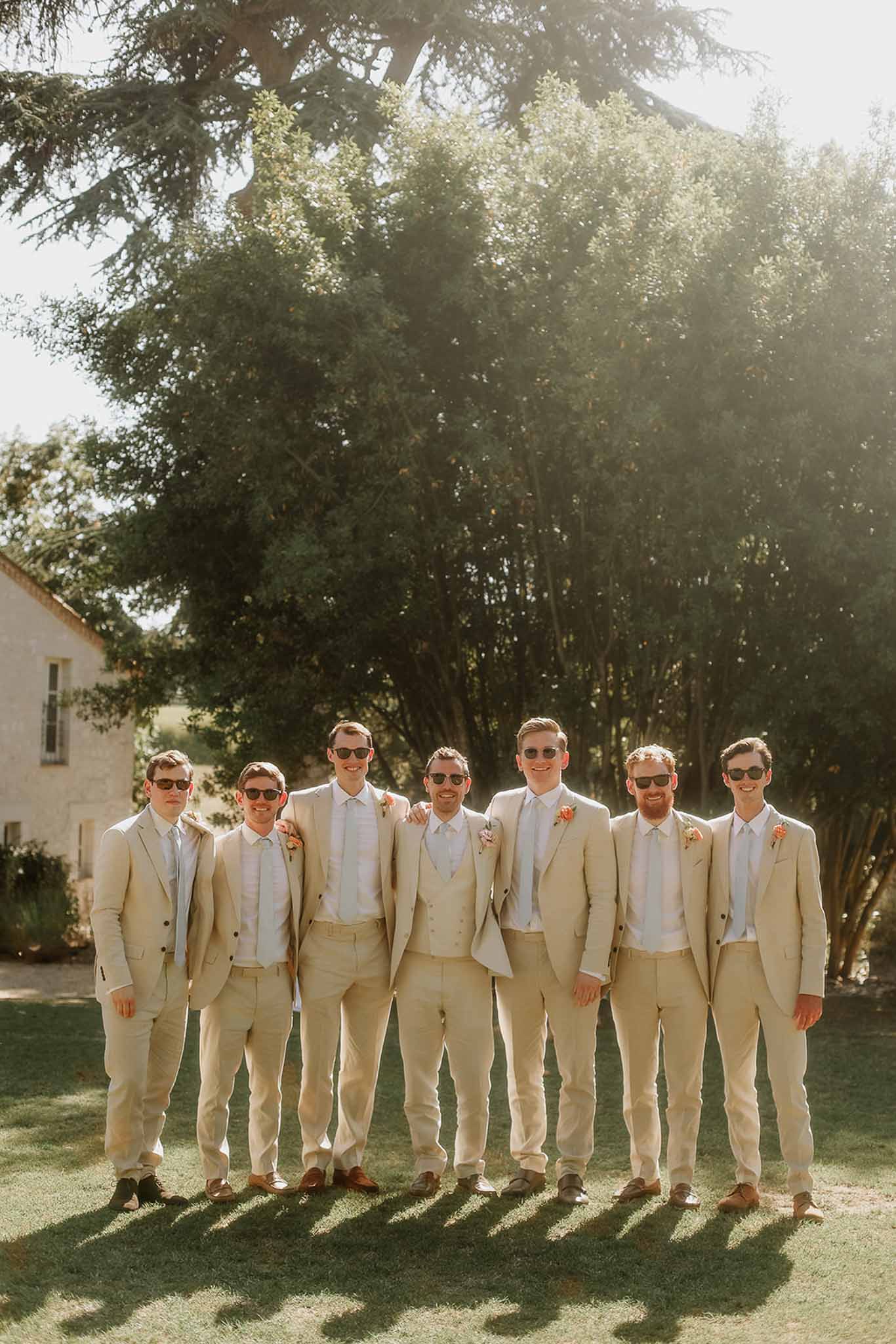 Groom and groomsmen in cream linen suits standing in line beneath trees at stone building