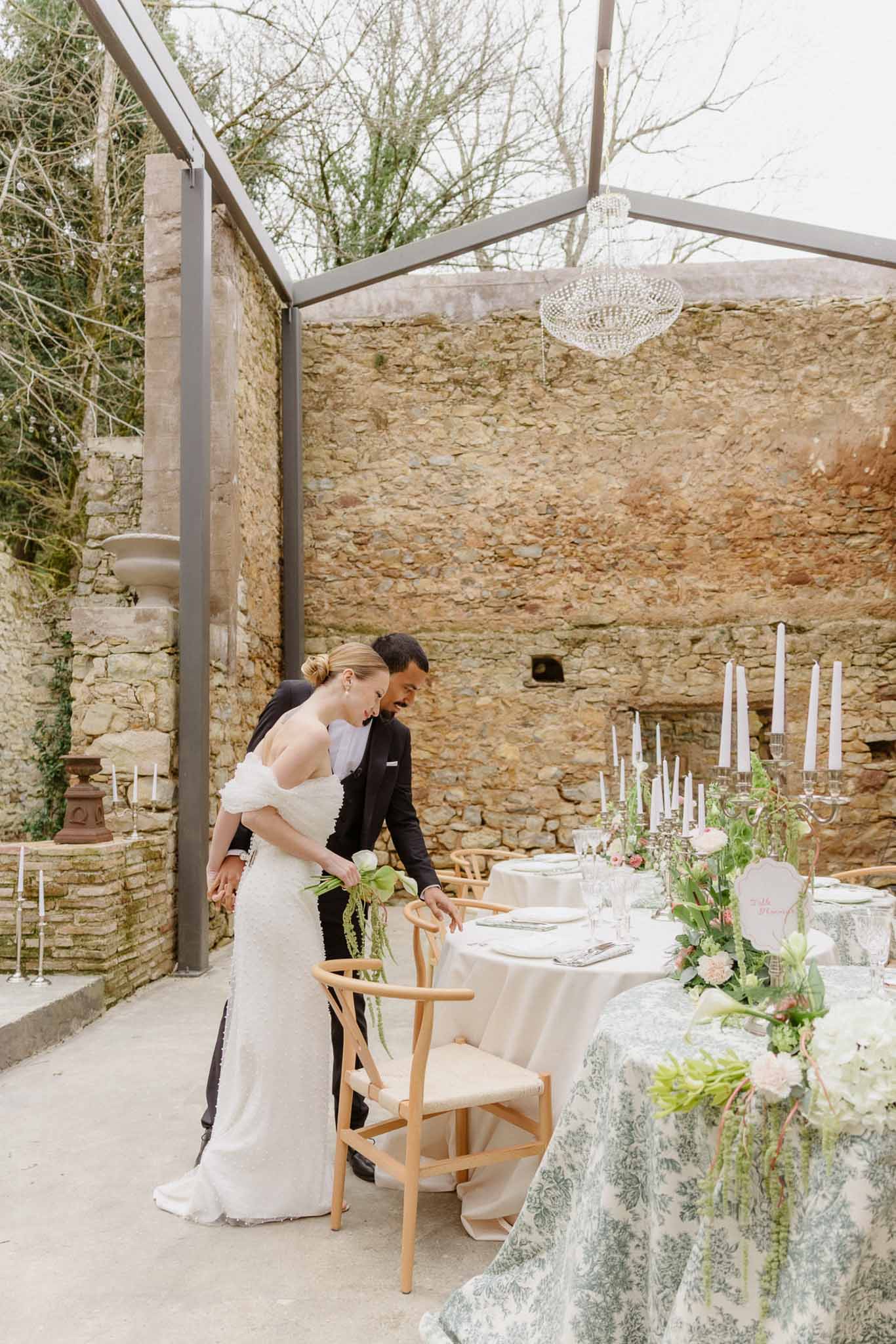 Bride and groom reviewing reception setup in stone courtyard with glass roof and floral table arrangements