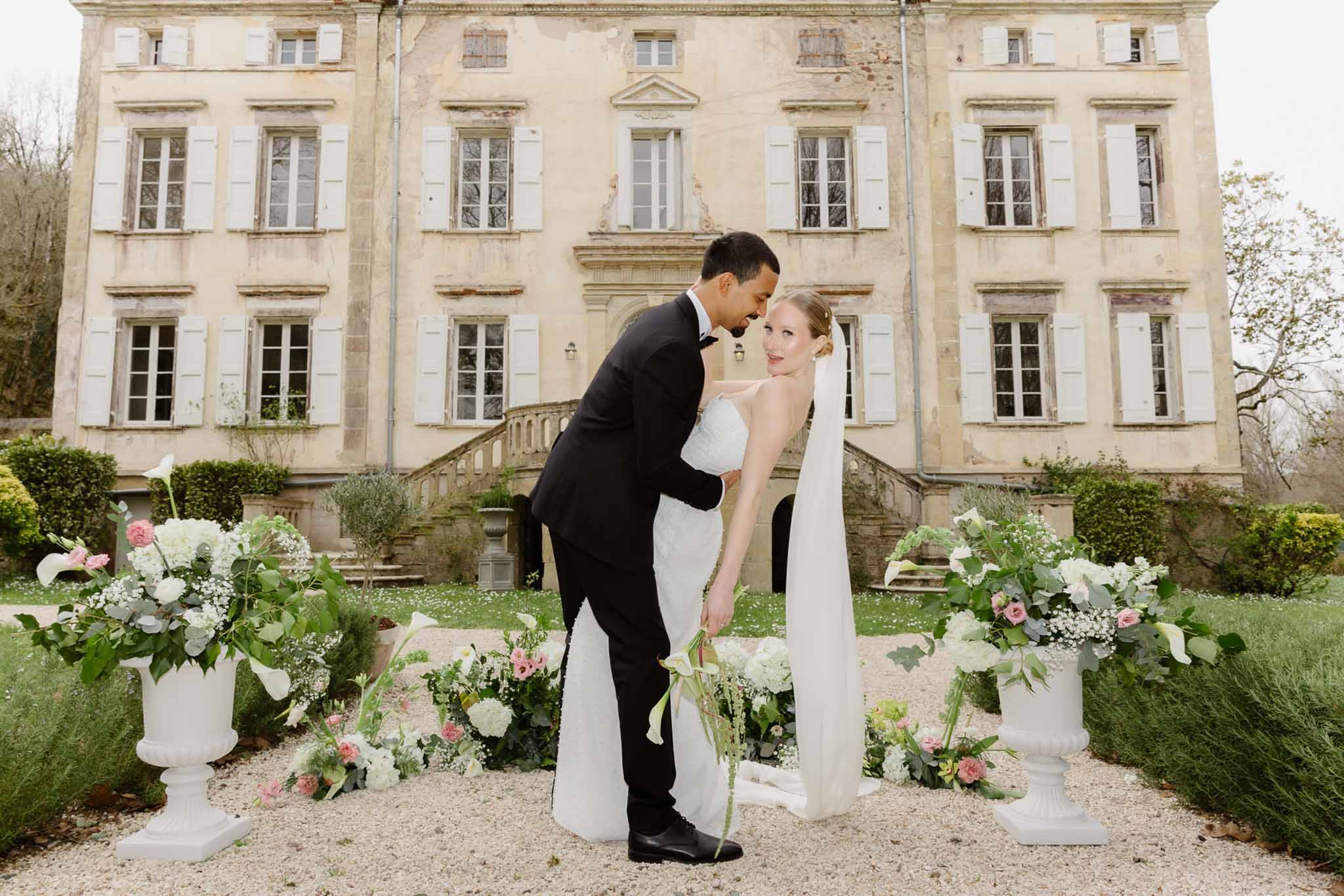 Bride and groom kissing in courtyard of French château with floral arrangements and classical architecture