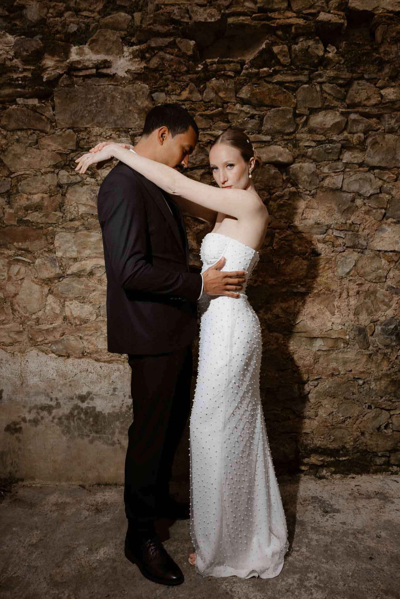 Bride in pearl-embellished strapless gown with groom leaning on her shoulder against stone wall
