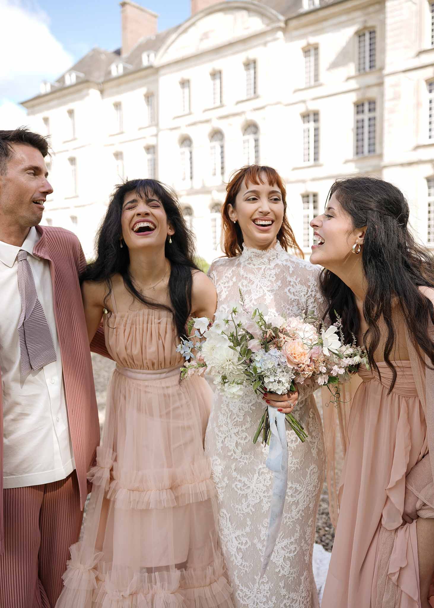 Bride and wedding party in dusty rose attire laughing in front of classical stone mansion