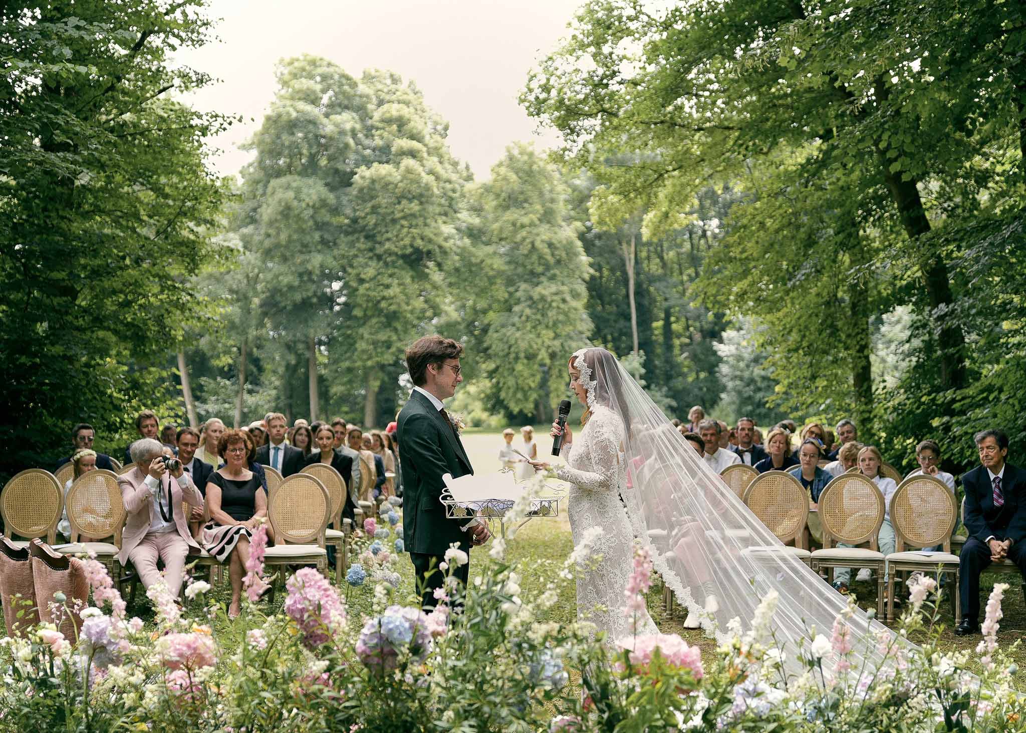 Outdoor garden wedding ceremony with bride and groom exchanging vows surrounded by seated guests in tree-lined allee