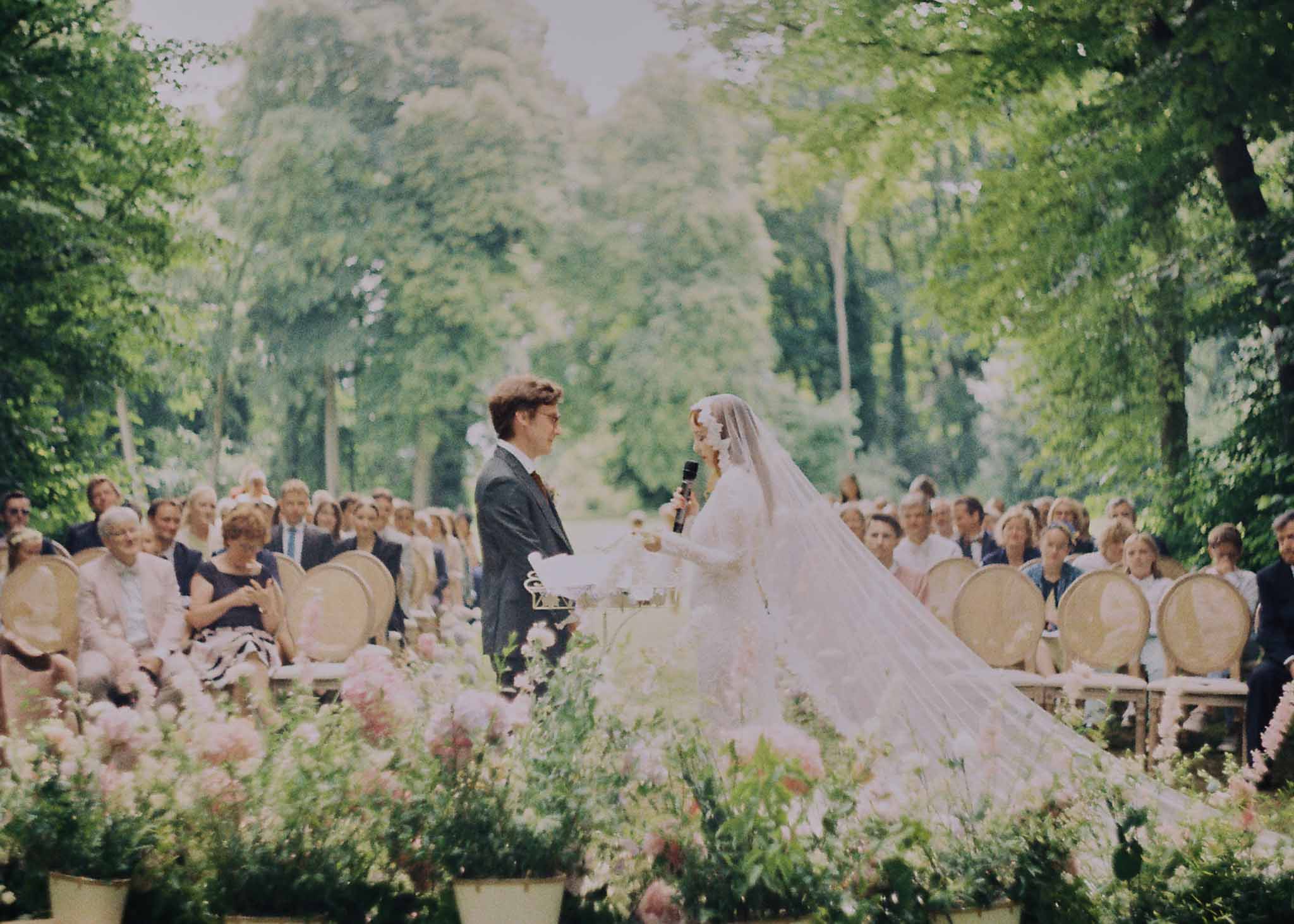 Outdoor wedding ceremony with bride and groom exchanging vows in formal garden with cypress trees and seated guests