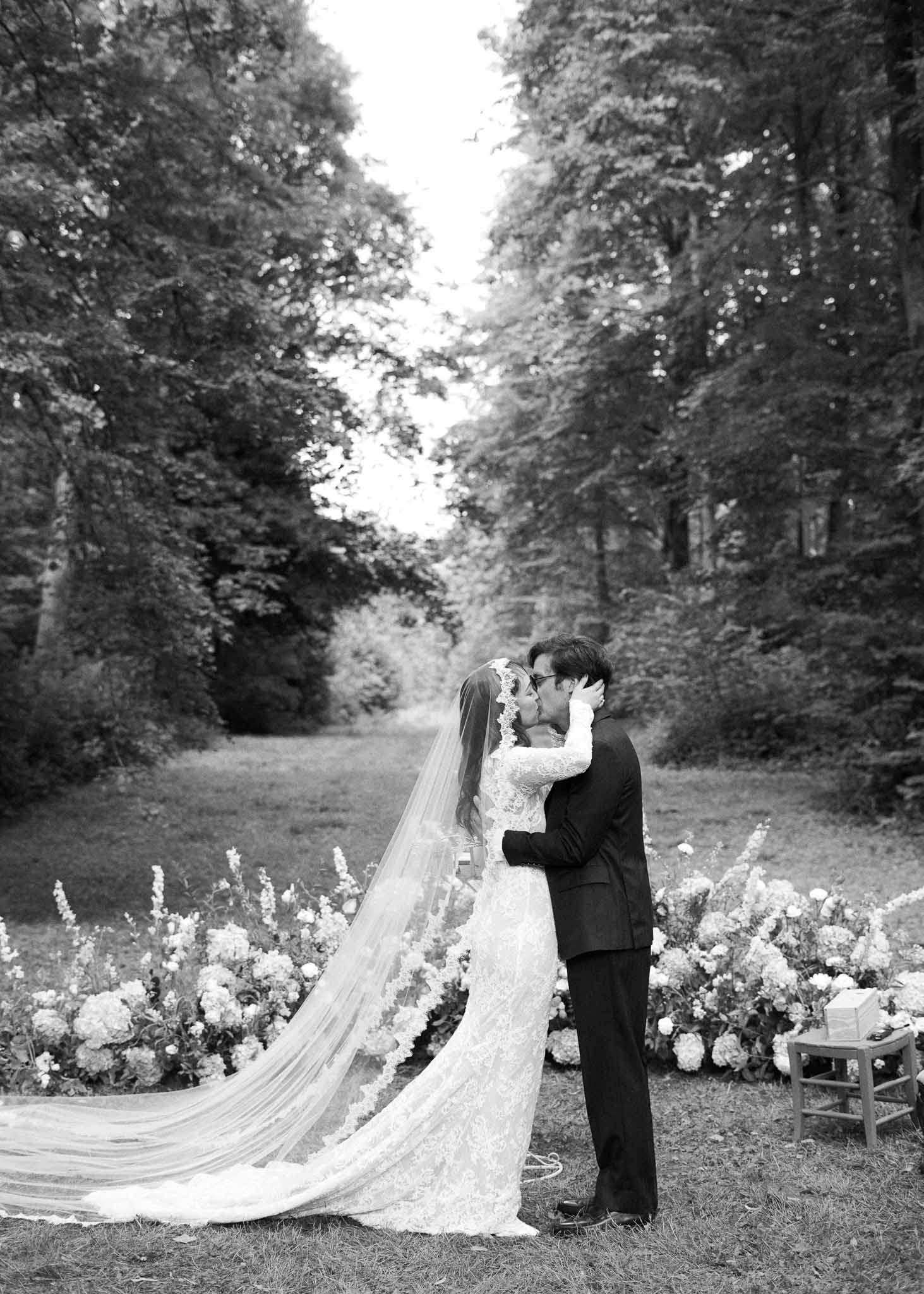 Bride and groom sharing first kiss in formal garden with white flowers and hedge-lined pathway