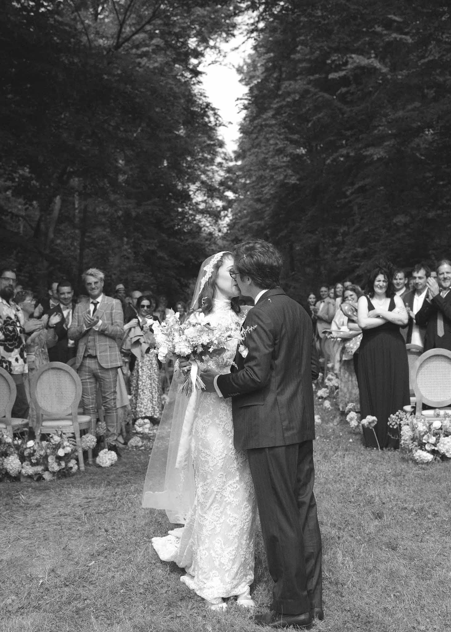 Bride and groom kissing at outdoor ceremony altar with guests watching in tree-lined garden setting